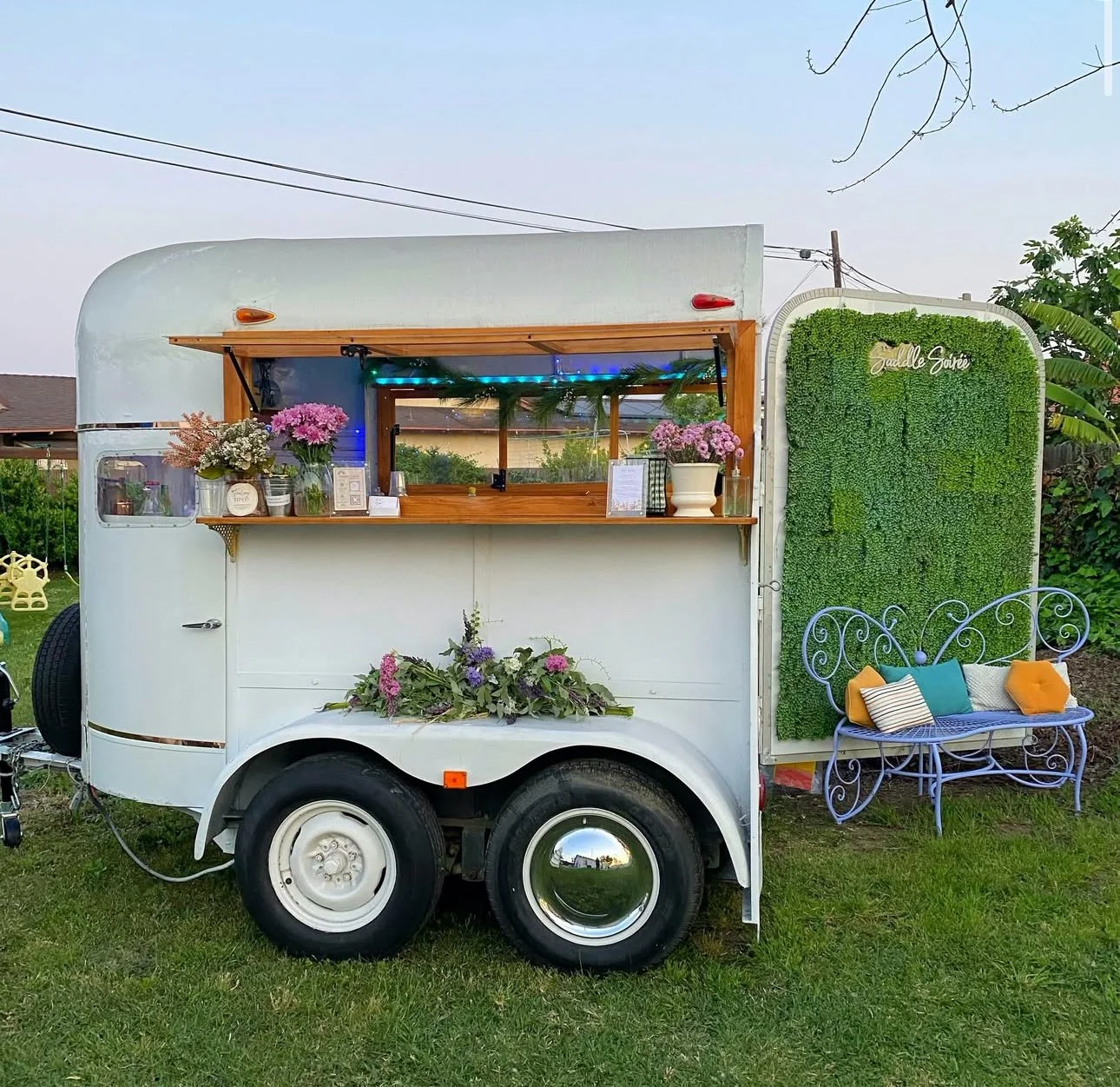 White vintage trailer decorated as a café with flowers, wooden counter, and colorful pillows on a blue metal bench outside. Green wall with sign, parked on grass yard.