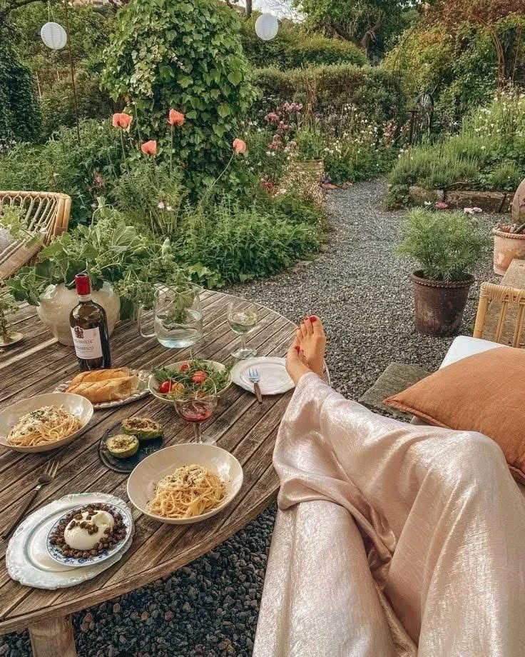 A volunteer's feet with red nail polish resting on a couch next to a wooden table set for a meal outdoors in a garden. The table has pasta, salad, a bottle of wine, and a dessert.