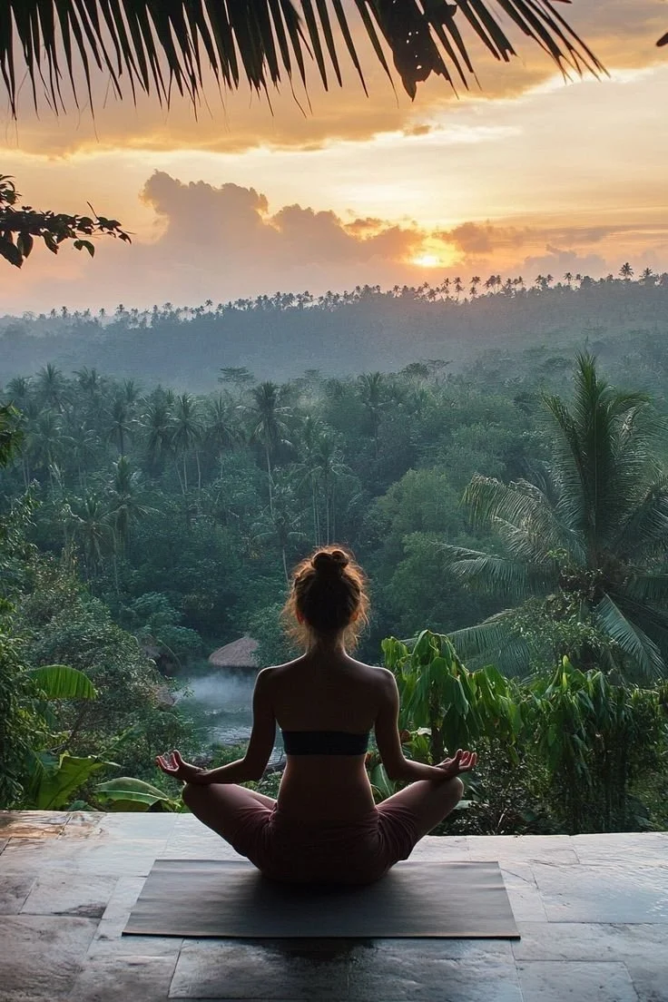 A woman practicing yoga in a seated meditation pose on a mat, overlooking a lush green jungle and a sunset sky.