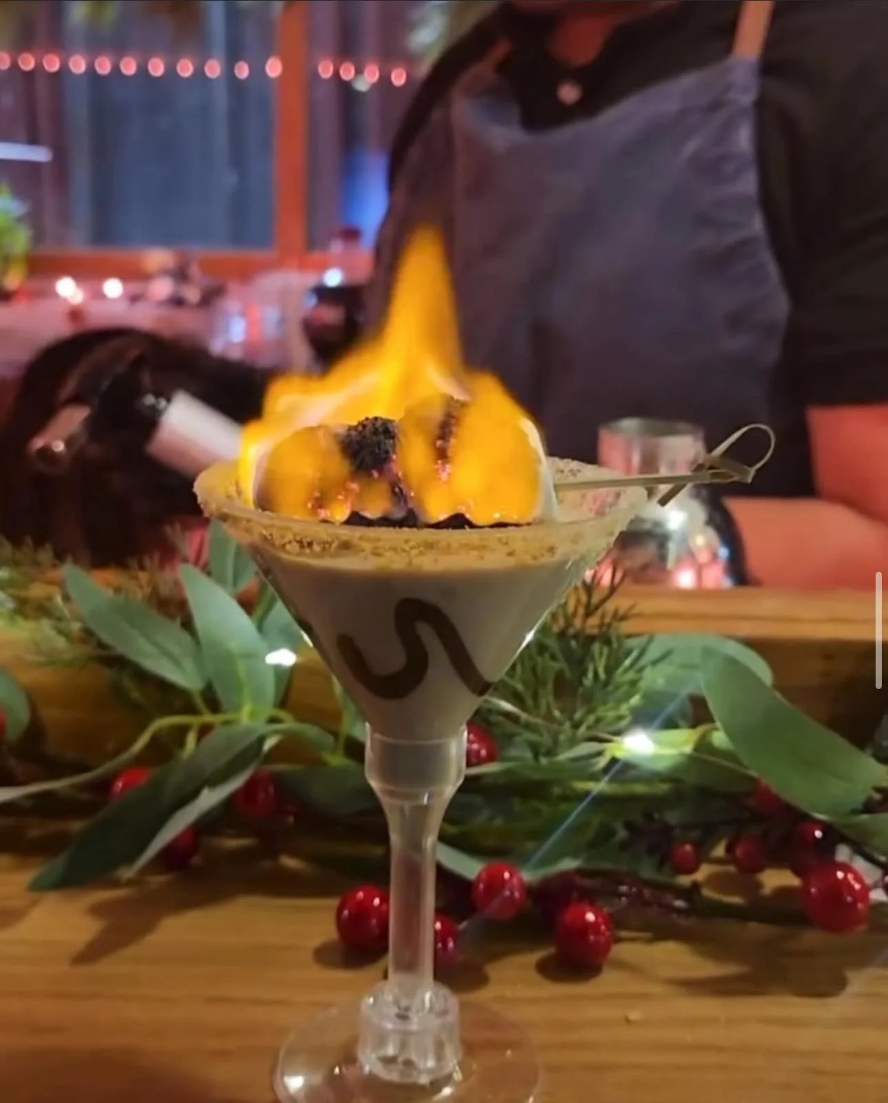 A dessert cocktail topped with a flame, with a decorative swirl design on the glass, surrounded by holiday greenery and red berries.