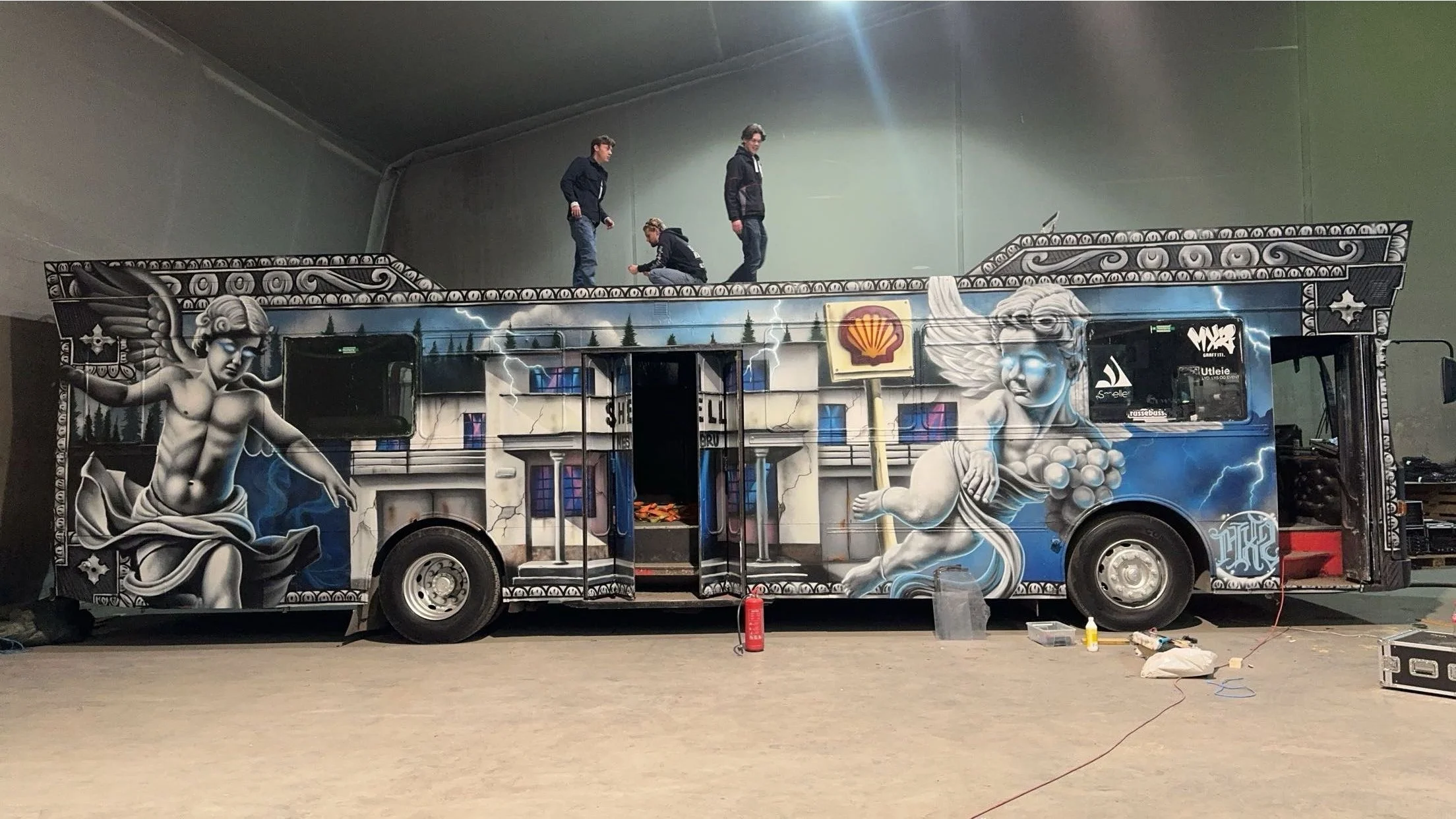 Et ombygd bus med graffiti-illustrasjon av to mytologiske gudinner, én med skjold og den andre med en drueklase, i et industrirom med tre personer som står på taket og jobber.