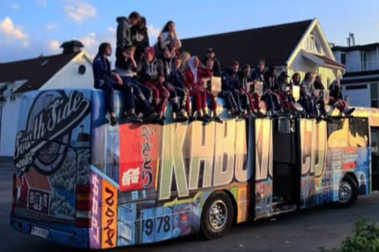 Mennesker sitter på taket av en dekorert bus, med en bolig i bakgrunnen.