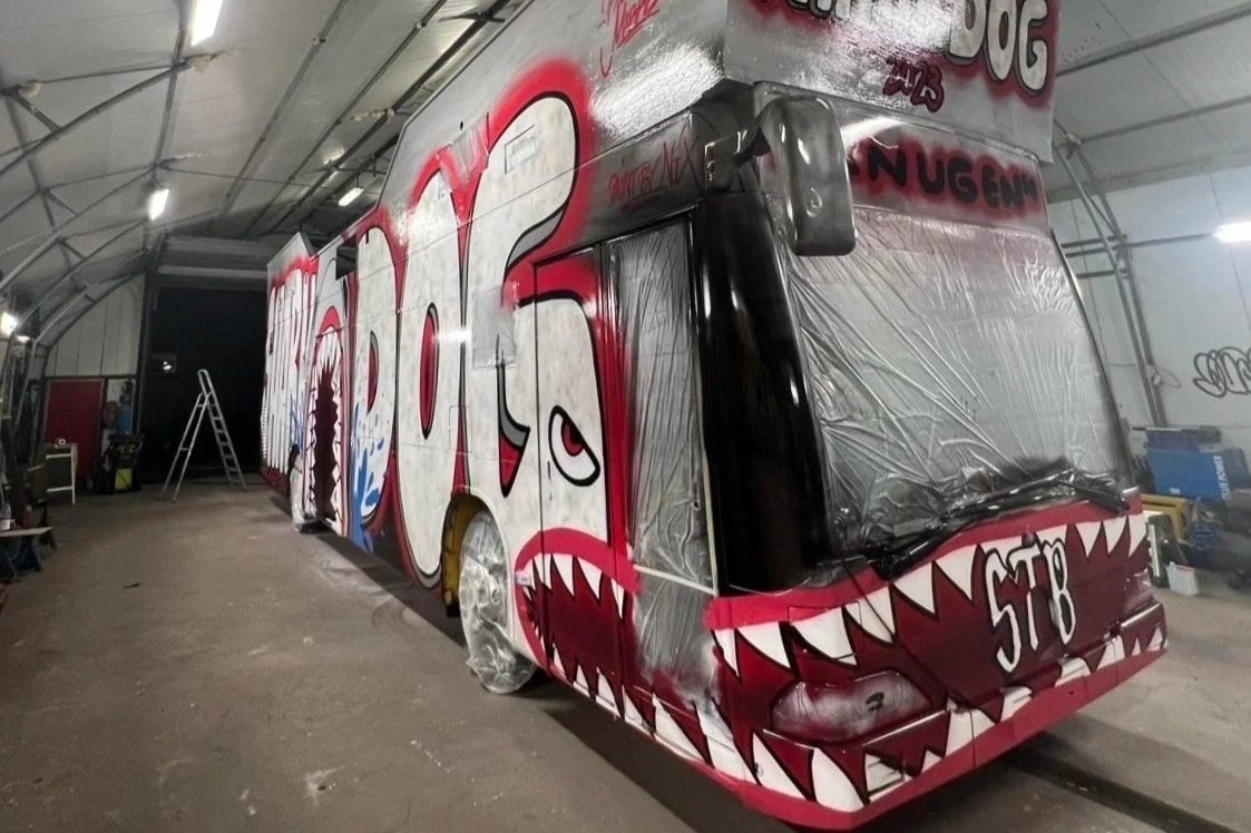 En enorm bus med graffiti og kunst på utsiden, som forestiller drageøyne og tenner, plassert innendørs i en lagerhallen.