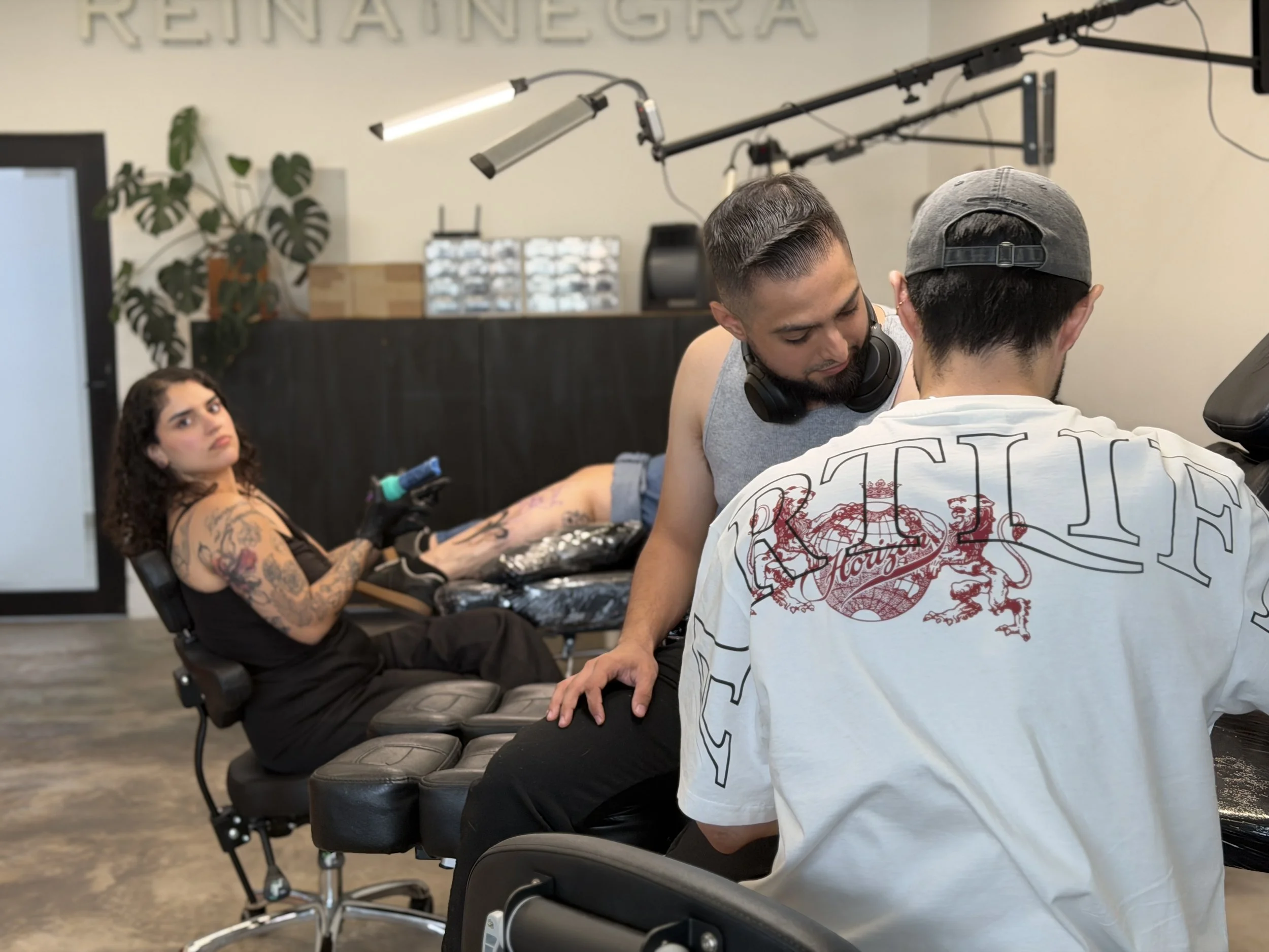 Una mujer con tatuajes en los brazos y cabello rizado sentado en una silla de tatuajes, con una expresión de atención y una aguja en mano. En el fondo, dos personas moretones, una con una camisa blanca con dibujos y la otra con auriculares y cerca de un equipo de tatuaje.