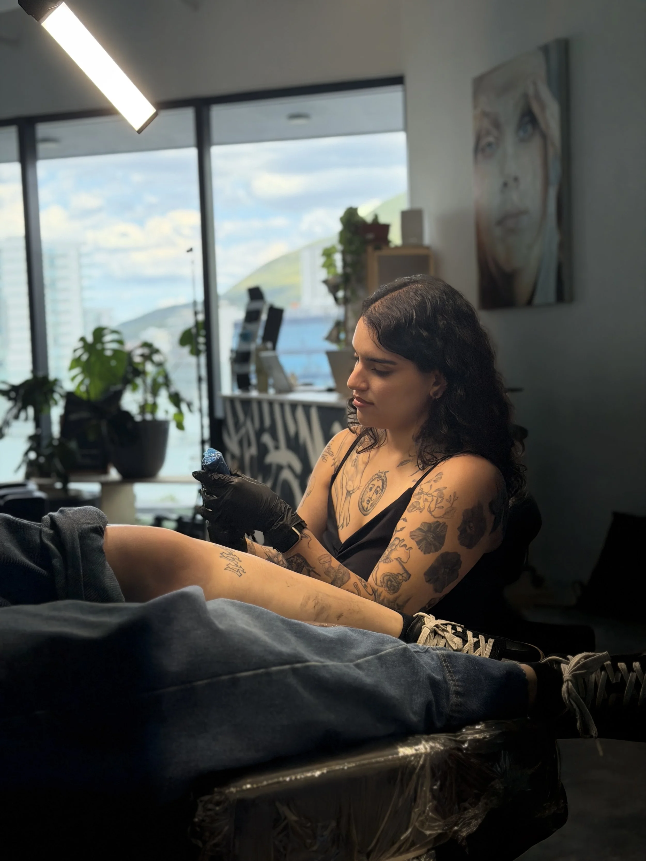 Mujer con tatuajes en brazos y piernas, sentada en una sala con ventanas grandes y vista a edificios y montañas, con un retrato de una mujer en la pared, usando guantes negros y sosteniendo un objeto.