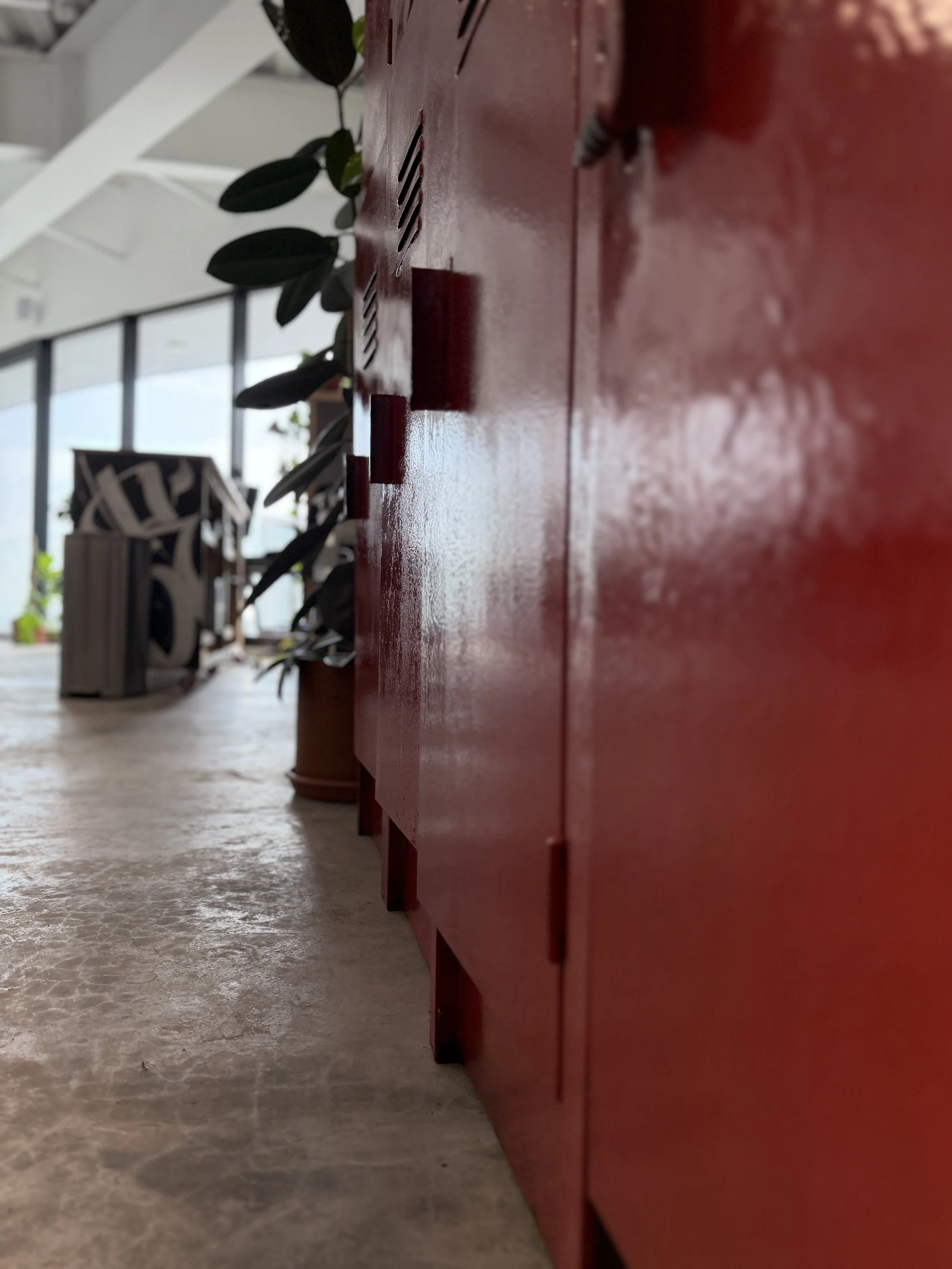 Vista lateral de un mueble rojo con ventilas, decorado con plantas verdes y una caja de almacenamiento de madera, en un ambiente bien iluminado y con ventanas grandes al fondo.