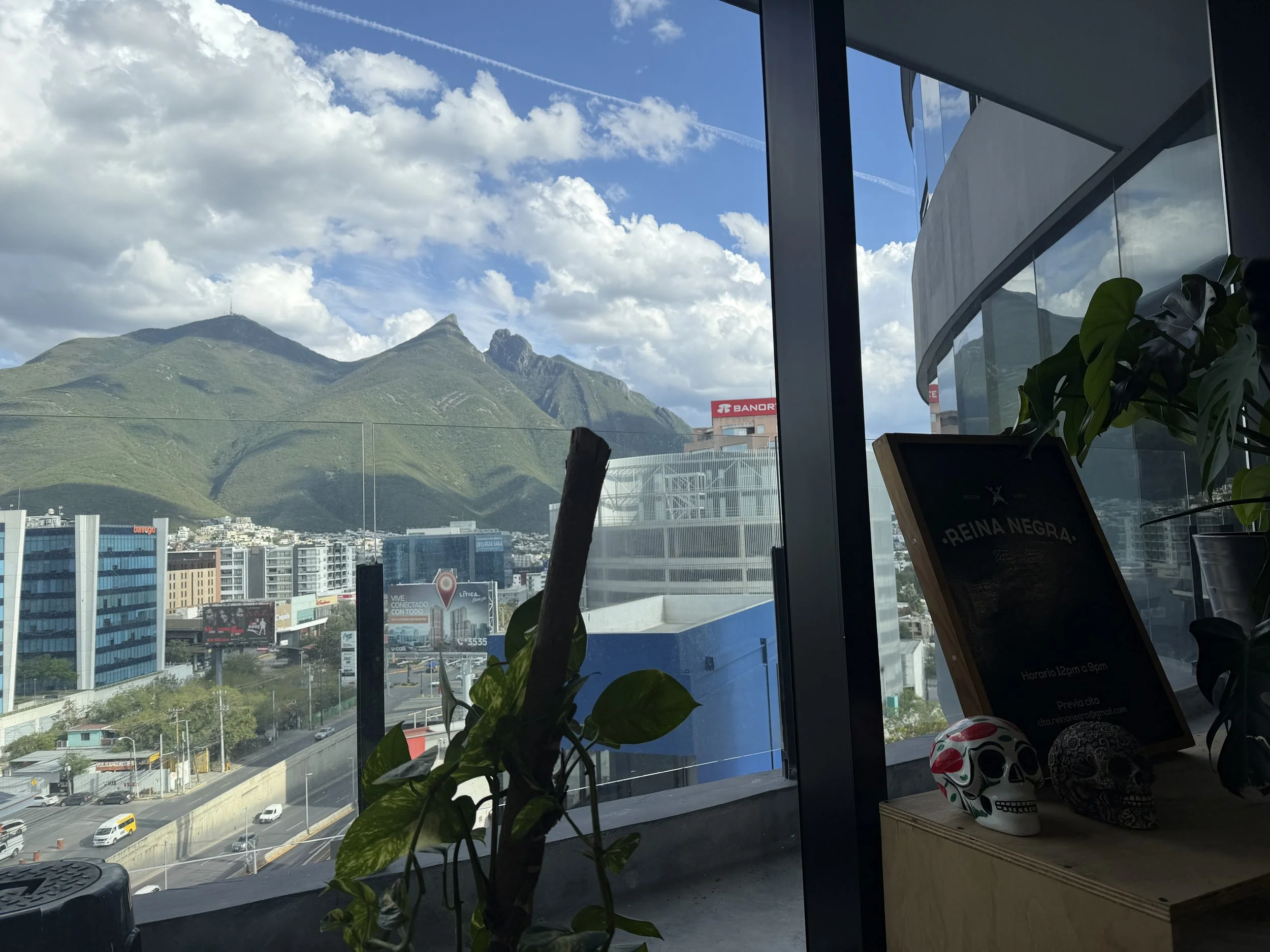Vista de un paisaje montañoso con cielo despejado y nubes, visto desde un interior de edificio con ventanas grandes, en una ciudad.