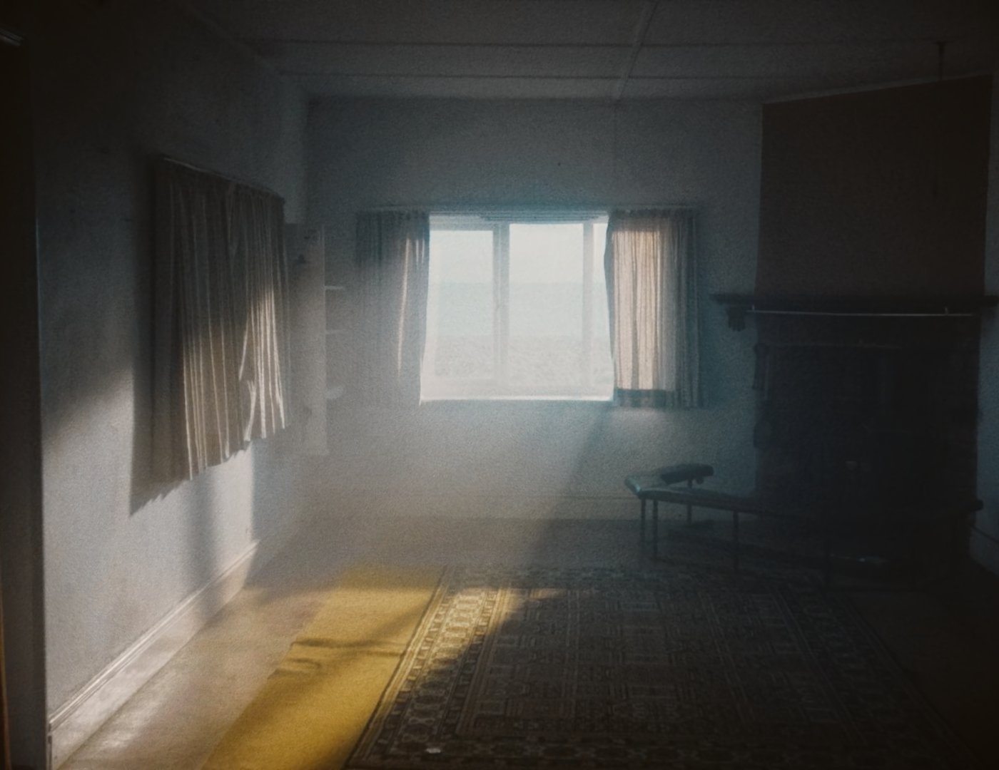 A dimly lit room with a window letting in natural light, old patterned rug, yellow runner, curtains, bench, and fireplace.