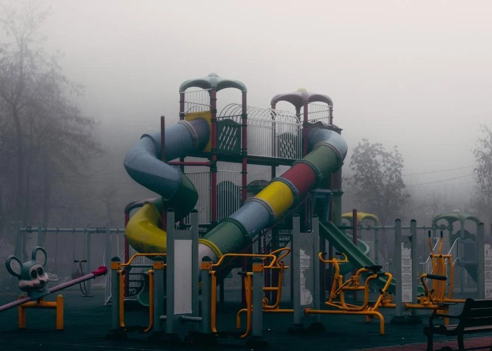 A foggy outdoor playground with a multi-colored tube slide, exercise equipment, and a mouse-shaped rocking toy in a park.