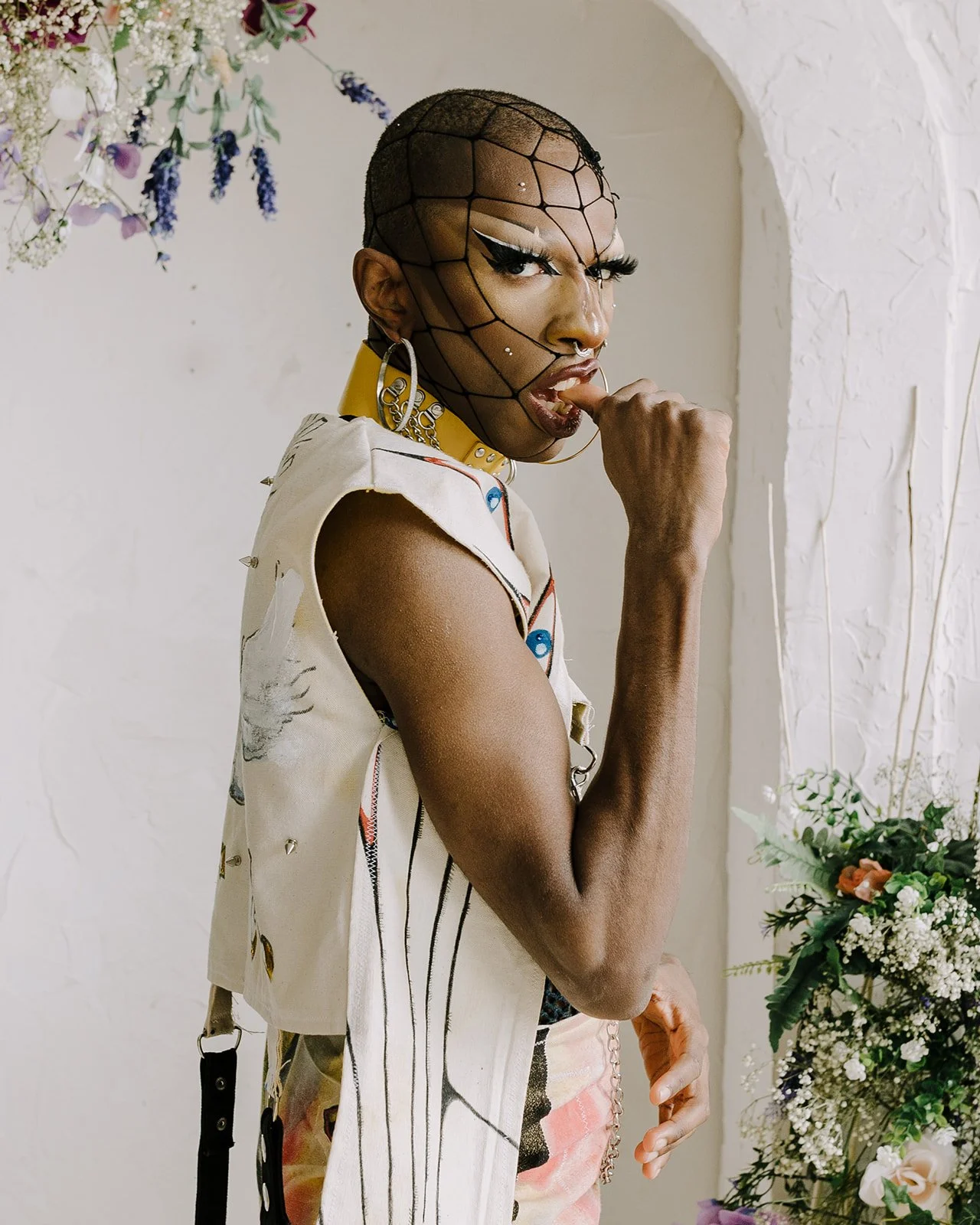 Person with bold makeup and a netted head accessory, wearing a sleeveless vest and patterned clothing, standing beside a floral arrangement