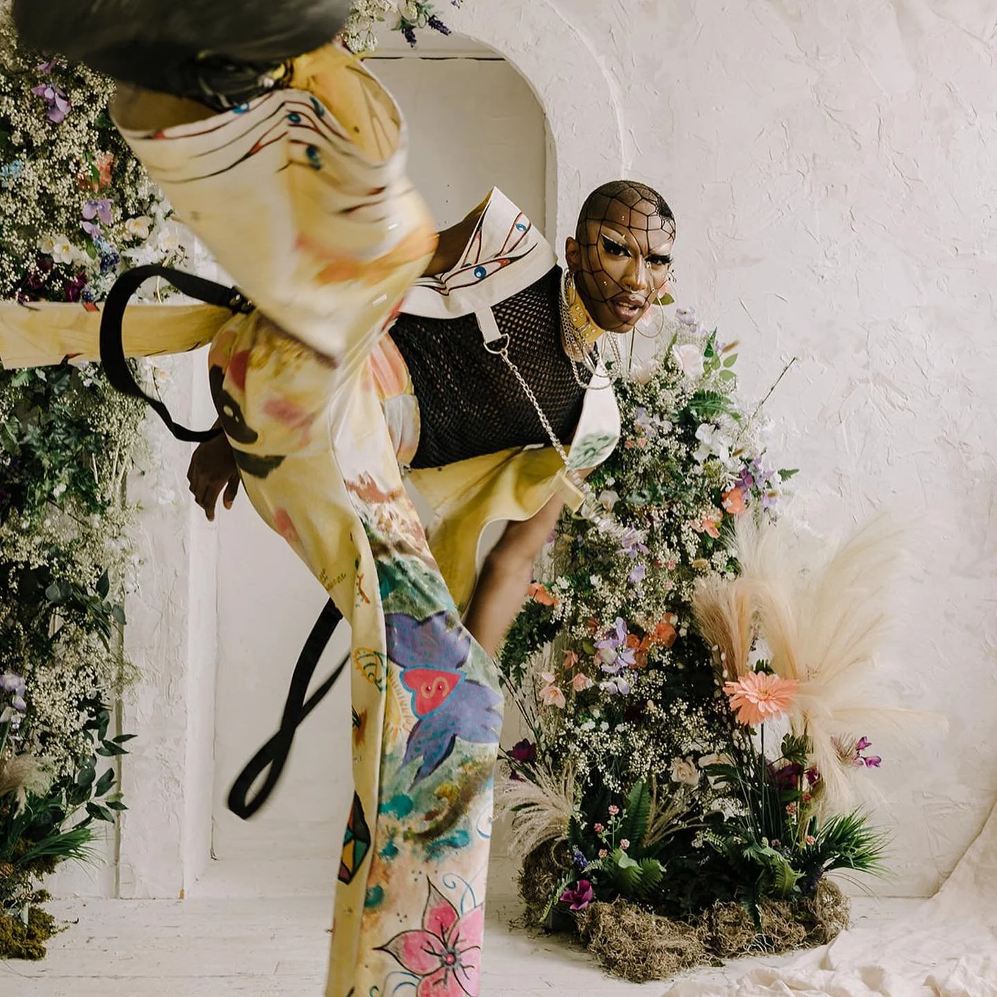 Person with bold makeup and jewelry, wearing vibrant, artistic clothing with floral and butterfly prints, posing among colorful flowers and greenery in an indoor setting.