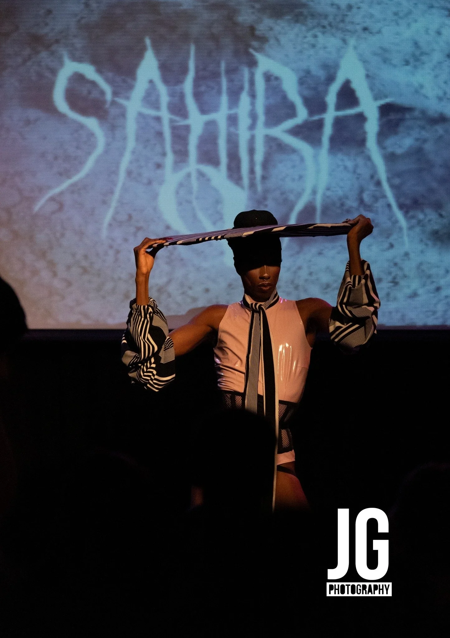 A woman on stage holding a striped scarf above her head with both hands. She is wearing a black head covering, a pink and black outfit, and striped sleeves. The background features a shadowy projection of the word 'SAHIRA' in stylized font, with the logo 'JC Photography' in the lower right corner.