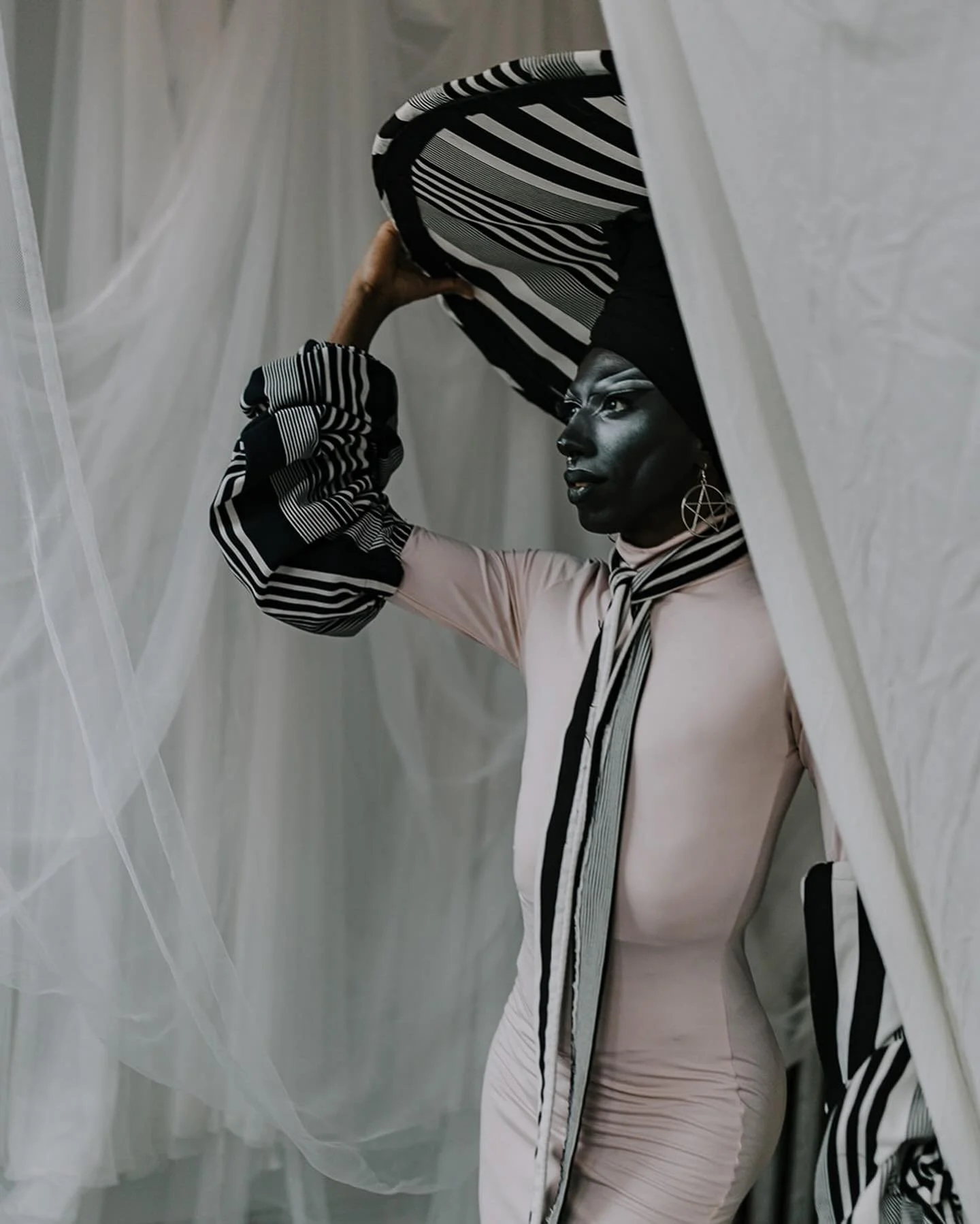 A person with dark skin and makeup, wearing large pentagram earrings and a pink outfit, holds a large striped fabric or hat overhead while standing behind sheer white curtains.