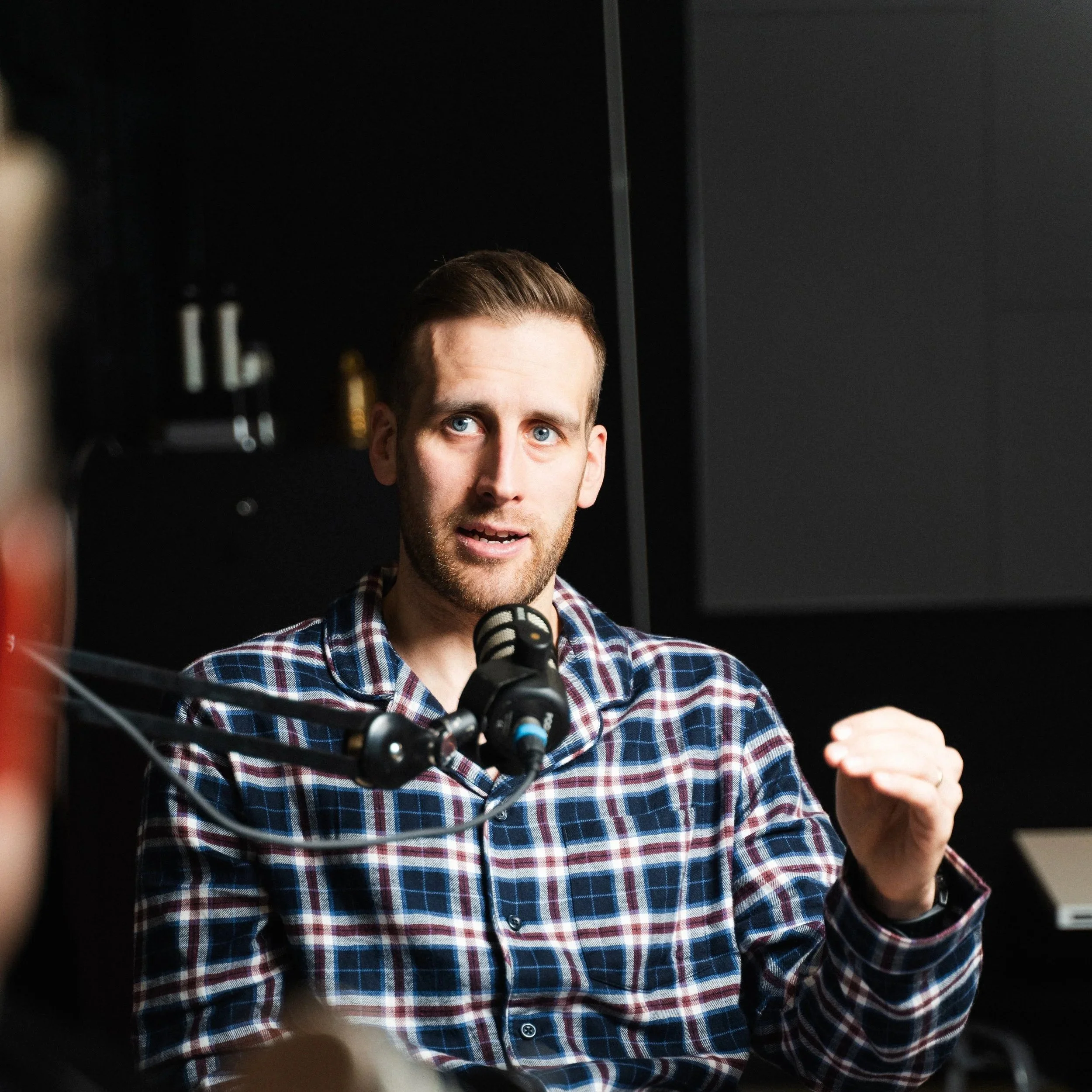 A man with blue eyes and brown hair speaking into a microphone in a dark studio setting, wearing a plaid shirt.