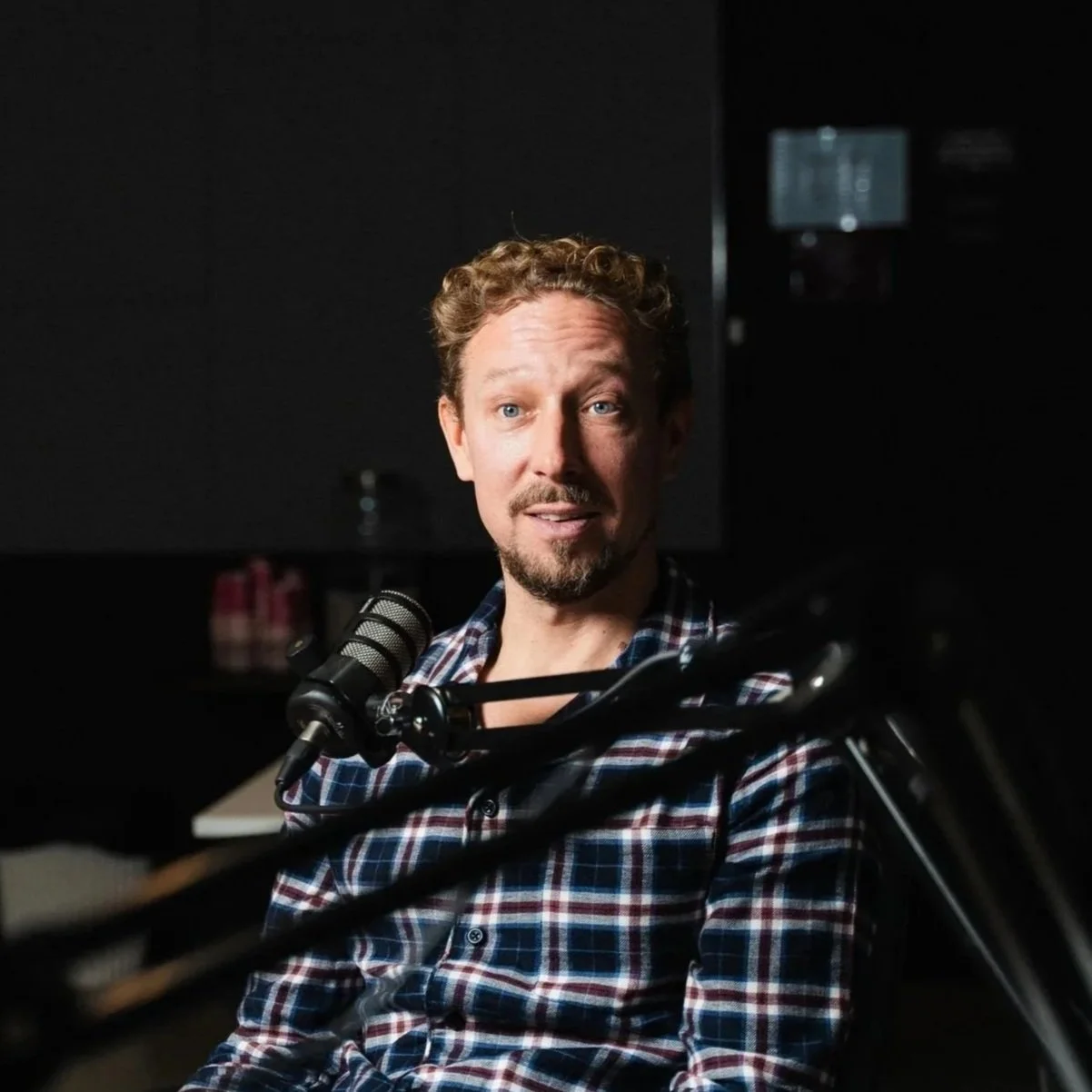 A man with curly hair and a beard speaking into a microphone in a dark room.