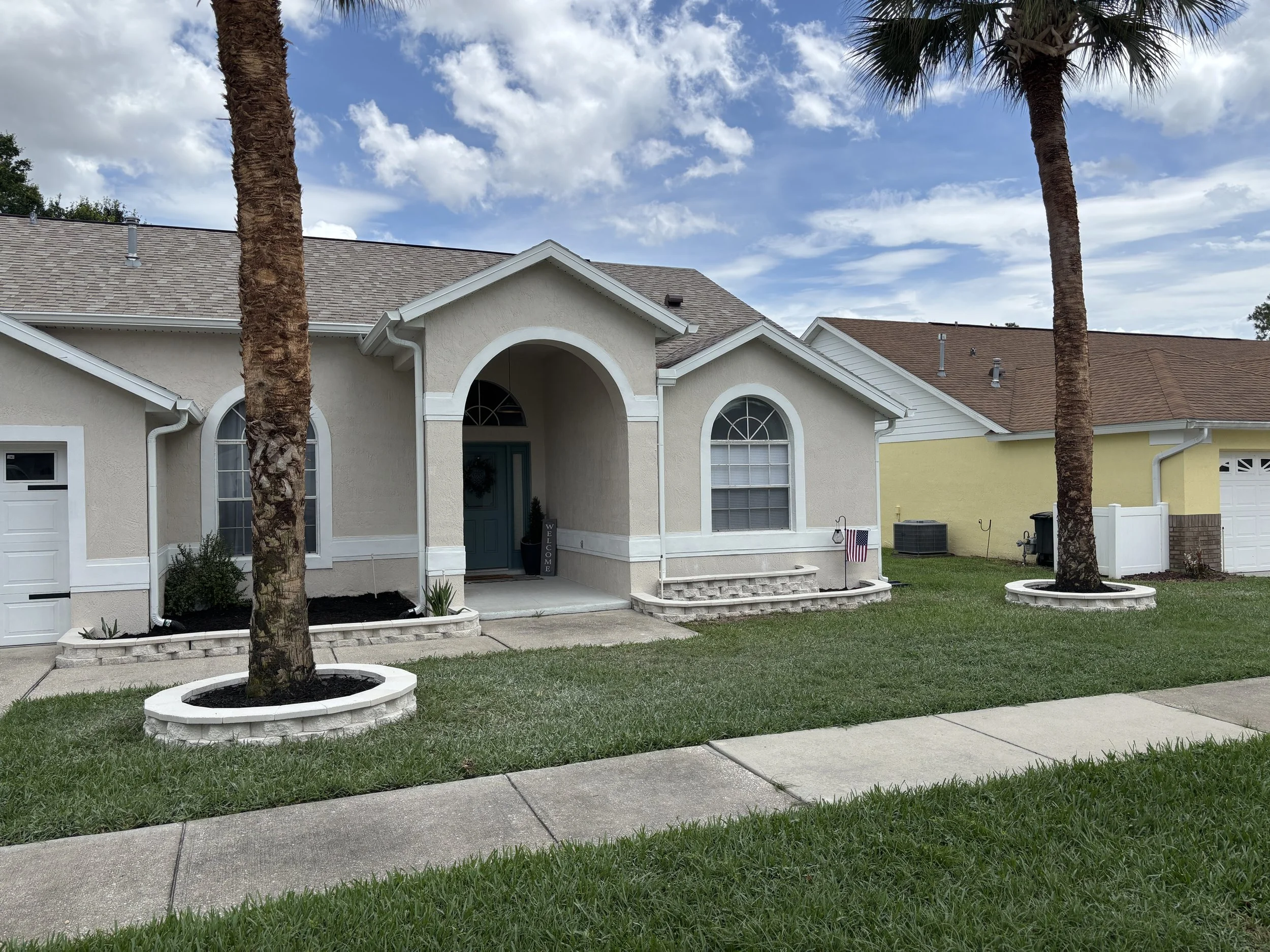 Two palm trees in front of a beige house with a gray roof and white trim, under a partly cloudy sky with blue and white clouds.