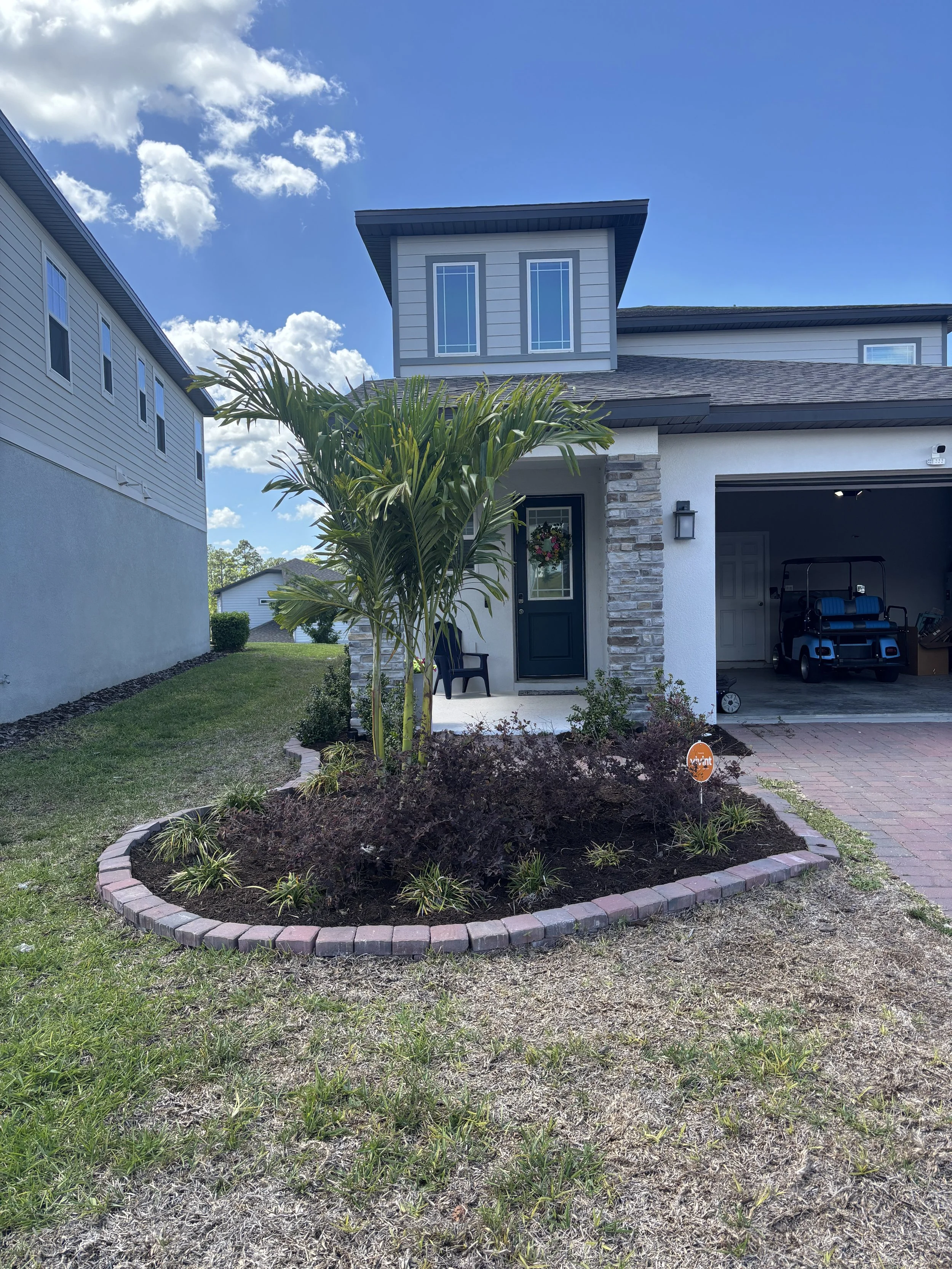 A landscaped front yard with a circular brick-edged garden bed containing plants and a small palm tree, grass, and a modern house with a garage and a golf cart parked inside. The house has a stone and white exterior, and the sky is partly cloudy.