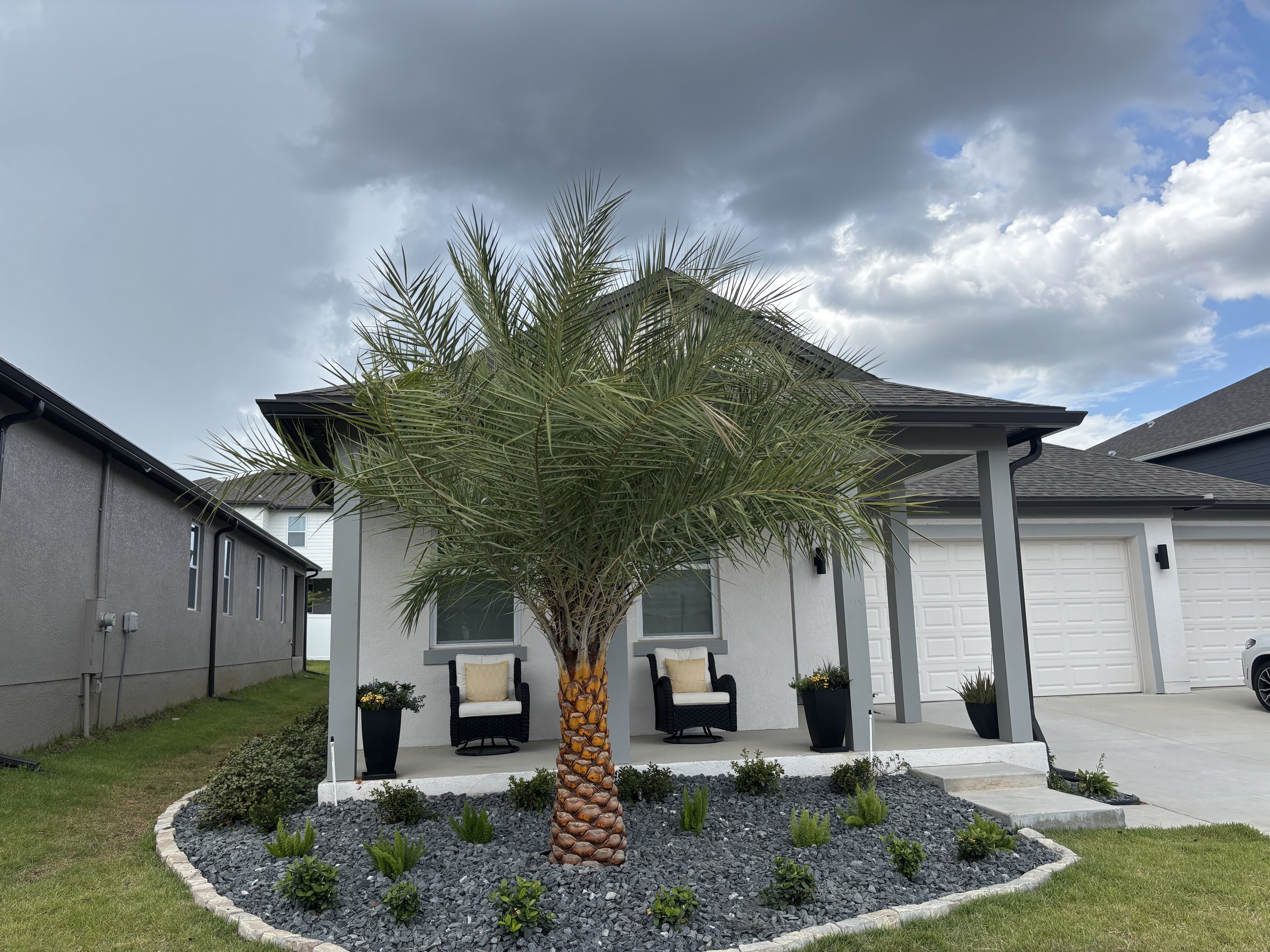 A palm tree in a landscaped front yard with a flower bed edged with bricks, two chairs with cushions, potted plants, and a gray house with a white garage door under a cloudy sky.