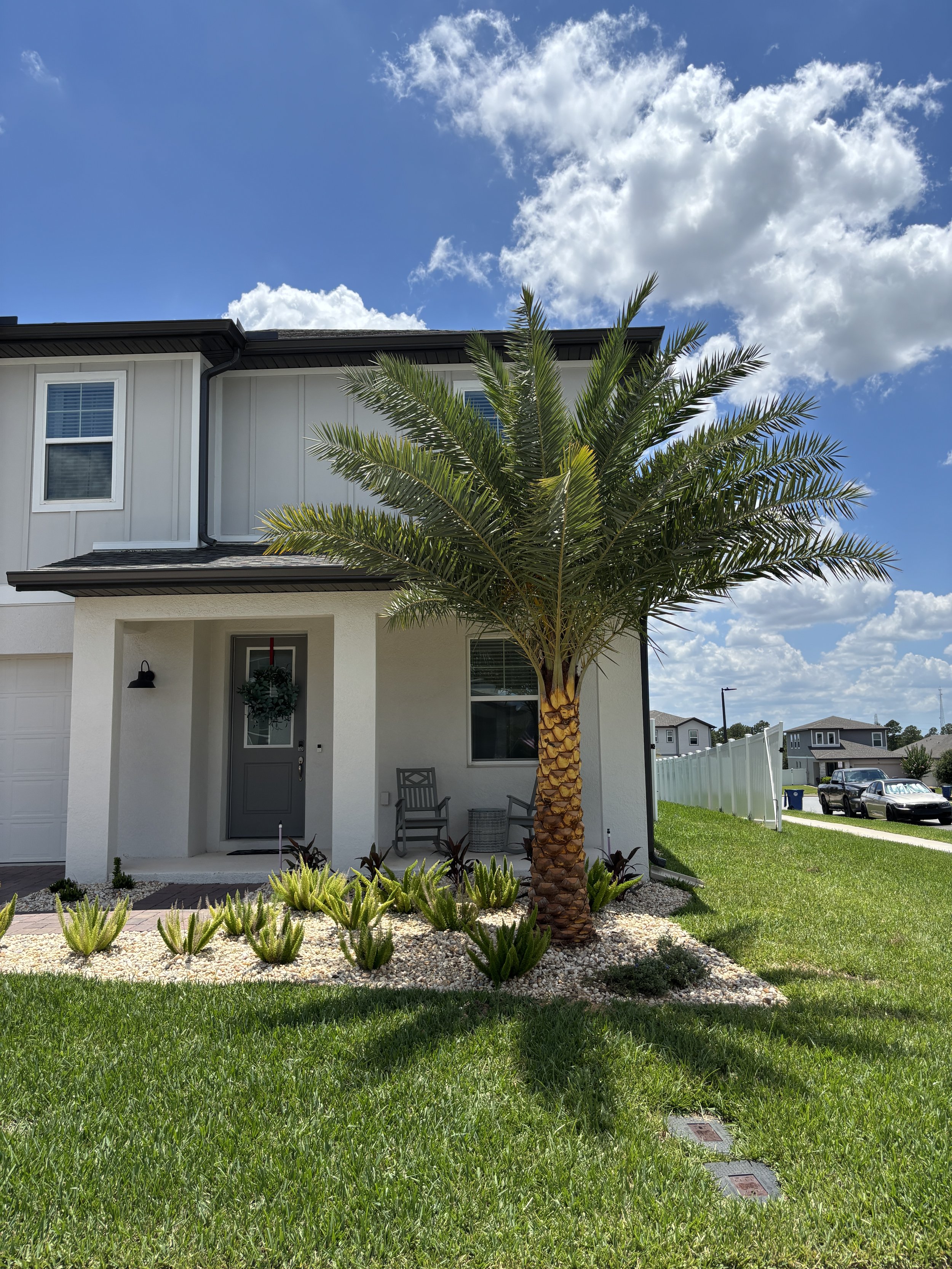 Front view of a modern house with a palm tree and a well-maintained green lawn, blue sky with white clouds, cars parked in the driveway, and neighboring houses in the background.