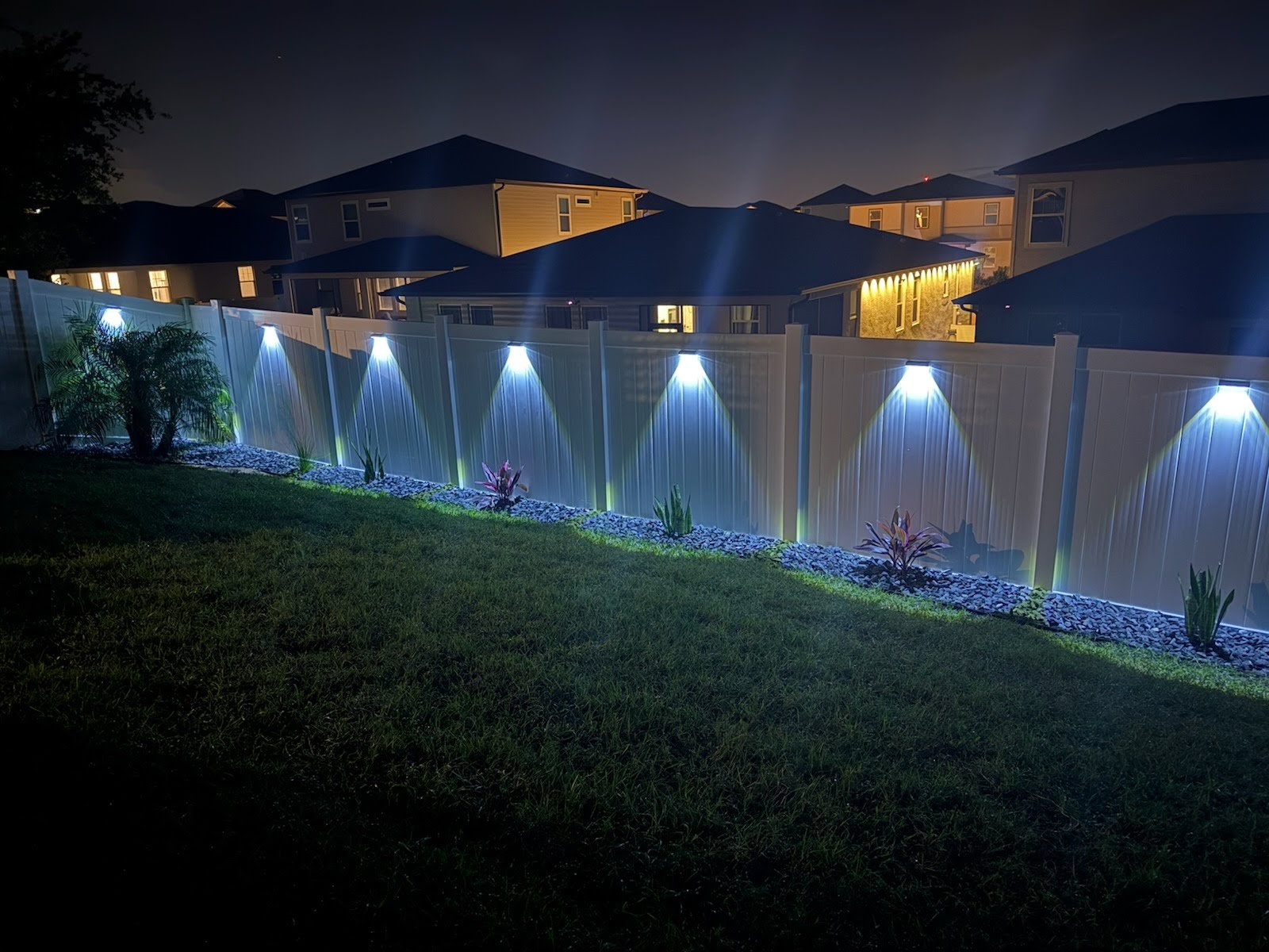 Nighttime view of a backyard with a white fence illuminated by blue outdoor lights, with a well-maintained lawn and small plants along the edge of the fence.