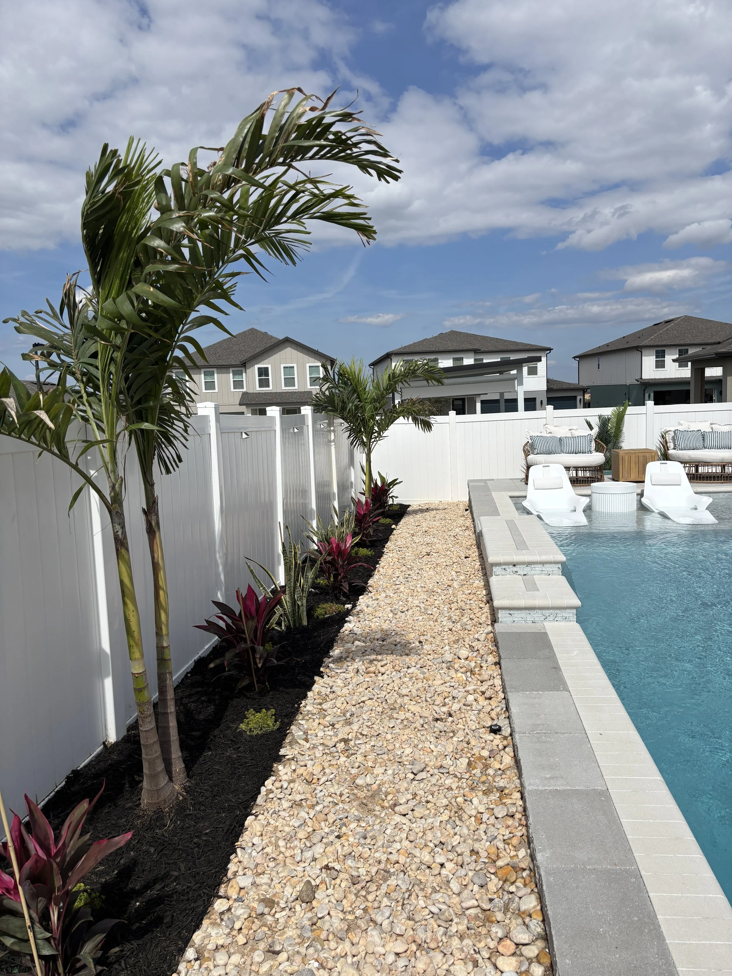 Backyard pool area with white lounge chairs, potted plants, a gravel walkway, painted houses, and a partly cloudy sky.