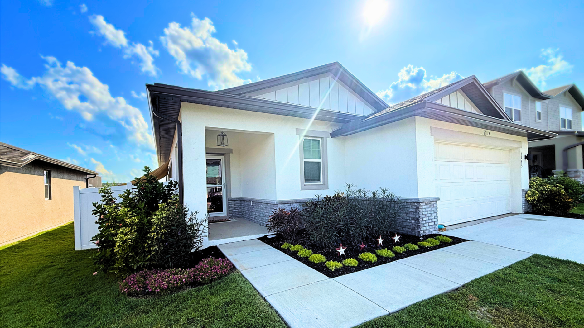 Modern white house with a stone accent wall, porch light, and a landscaped front yard with flowers and bushes, under a sunny blue sky.