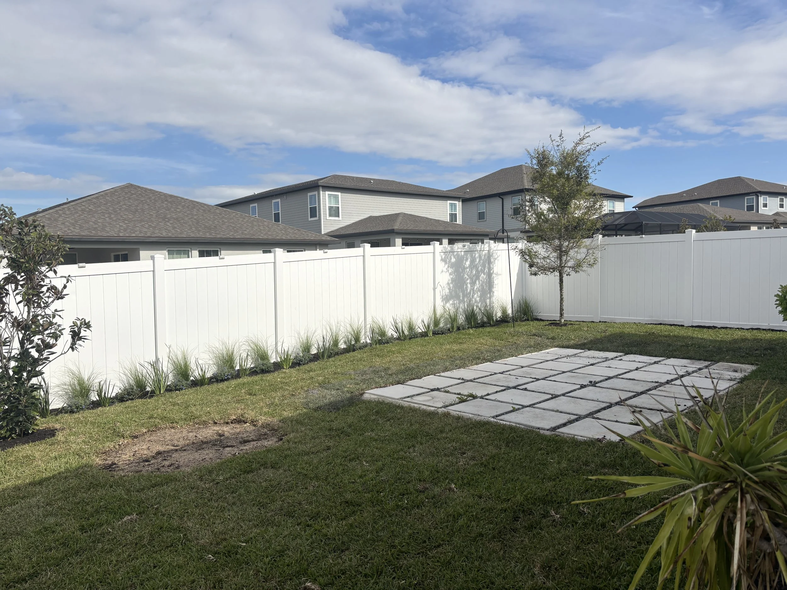 A backyard with a white fence, green grass, small trees, and paver stones, with houses and a blue sky with clouds in the background.