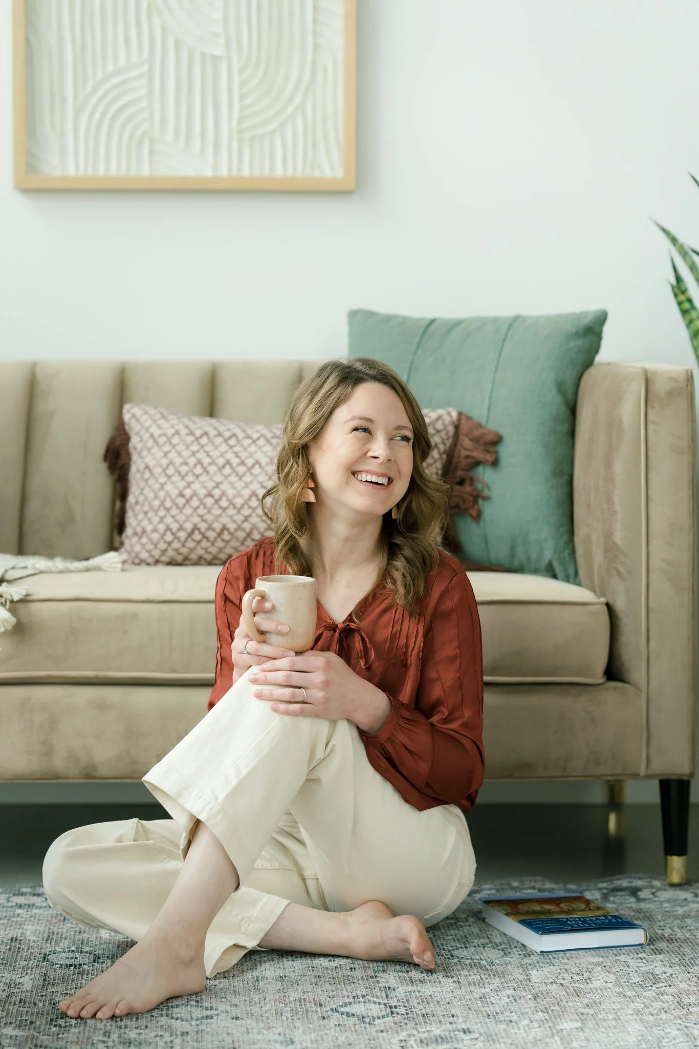 A woman sitting on the floor with her legs crossed, holding a mug, smiling, with a sofa and cushions in the background.