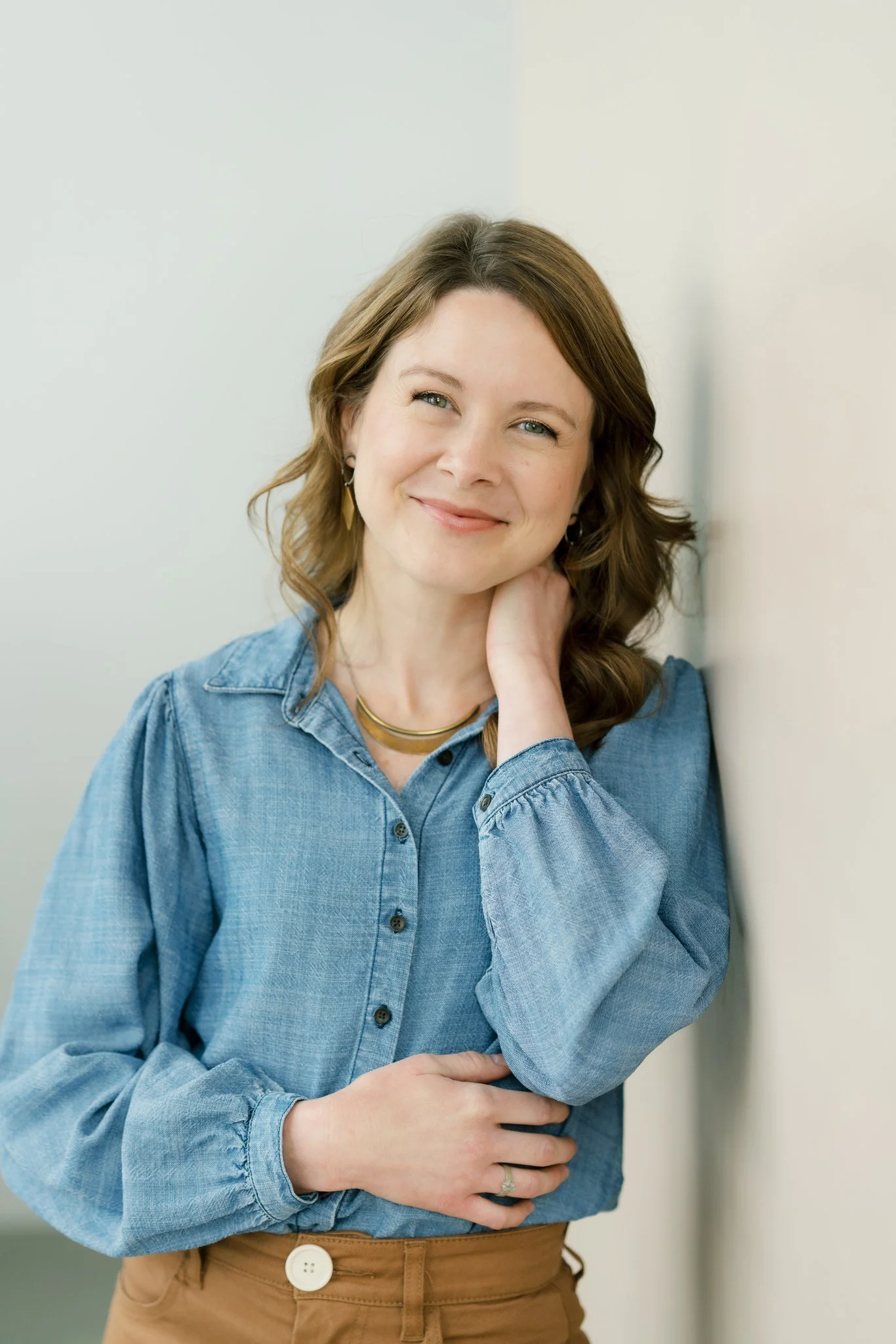 A woman with wavy brown hair, wearing a light blue denim shirt and tan pants, standing against an off-white wall, smiling softly, with her hand resting on her neck.