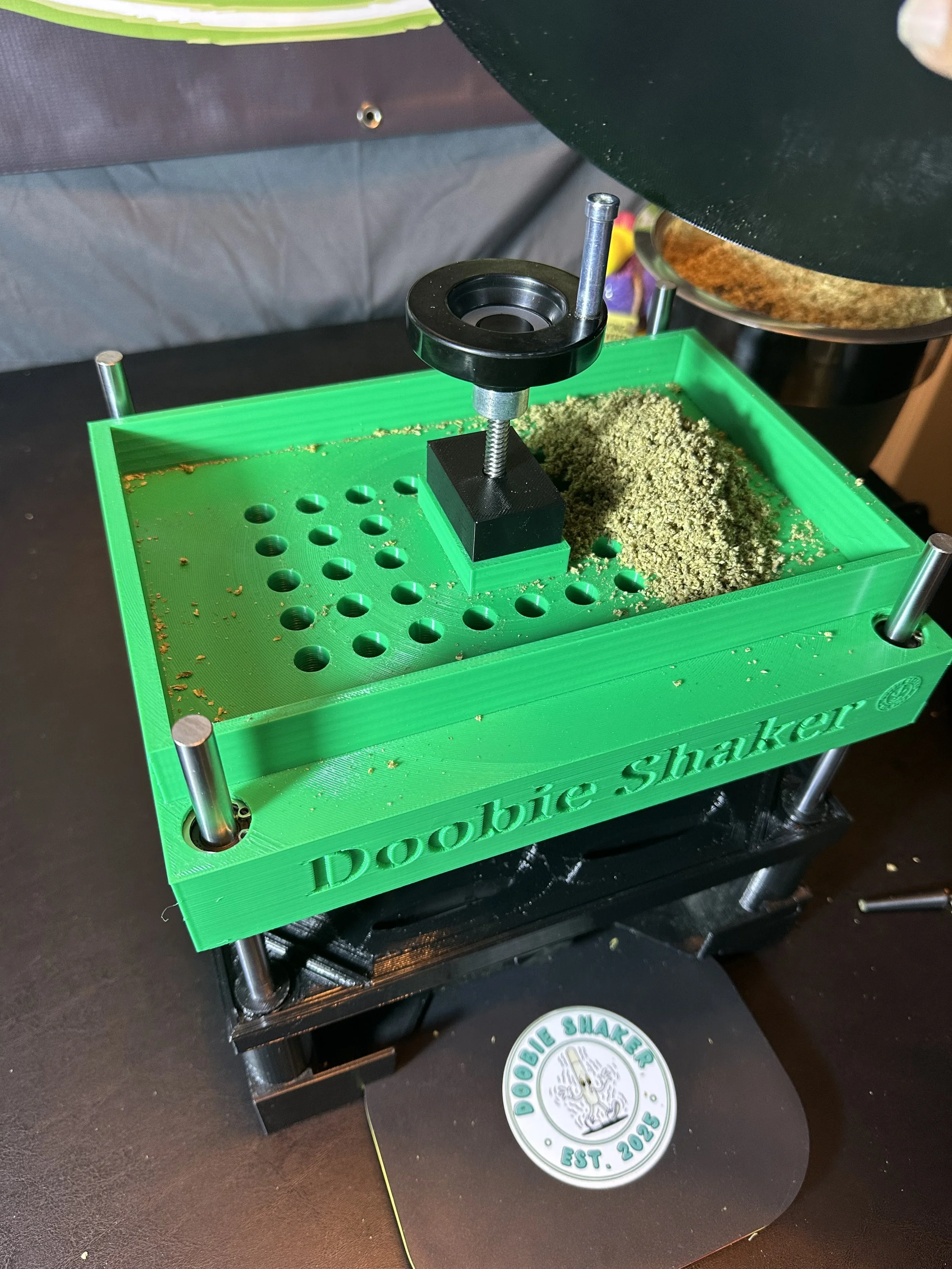 A green Doobie Shaker box with wooden frame and metal pegs, containing ground herbal material. A black and silver device with a circular component is positioned above the box, with supporting metal rods. The box has the words 'Doobie Shaker' engraved