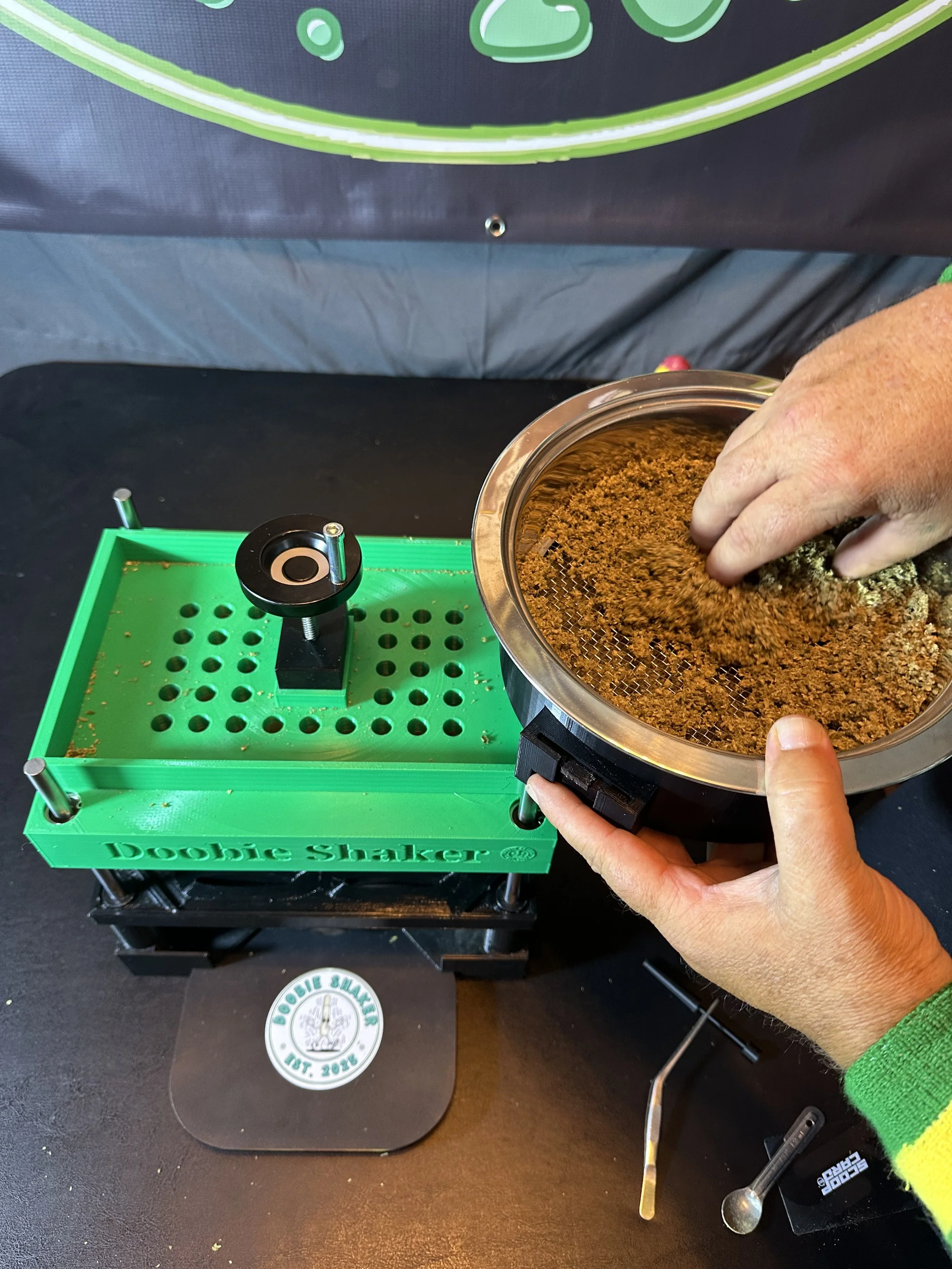 A person is using a Doobie Shaker to grind cannabis. The grinding process is in progress, with loose cannabis being directed into a container. The Doobie Shaker is a green and black device on a black table, with a person's hands managing the process.
