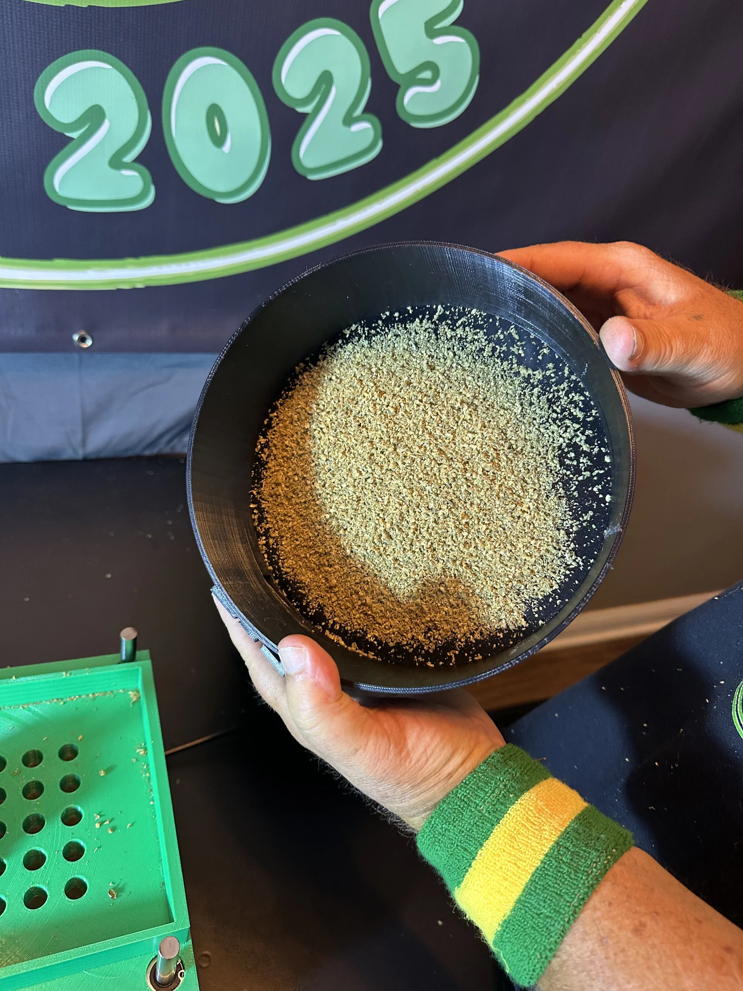 A person holding a black bowl containing yellowish-green granules, with a '2025' sign in the background.