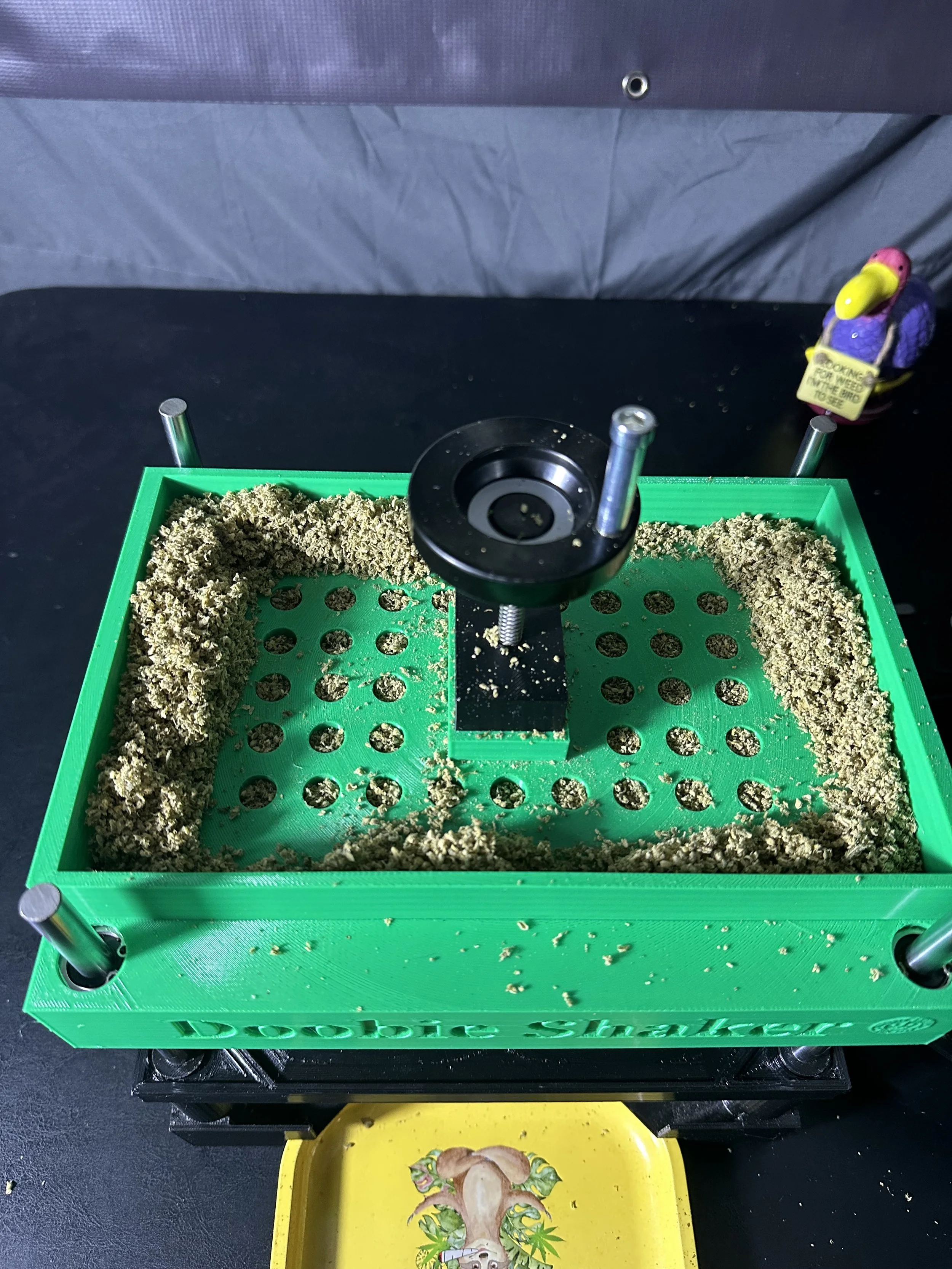 A green device with multiple holes and a central screw, surrounded by a beige textured material, on a black surface with a yellow tray beneath it. There is a bird figurine in the background holding a small sign.