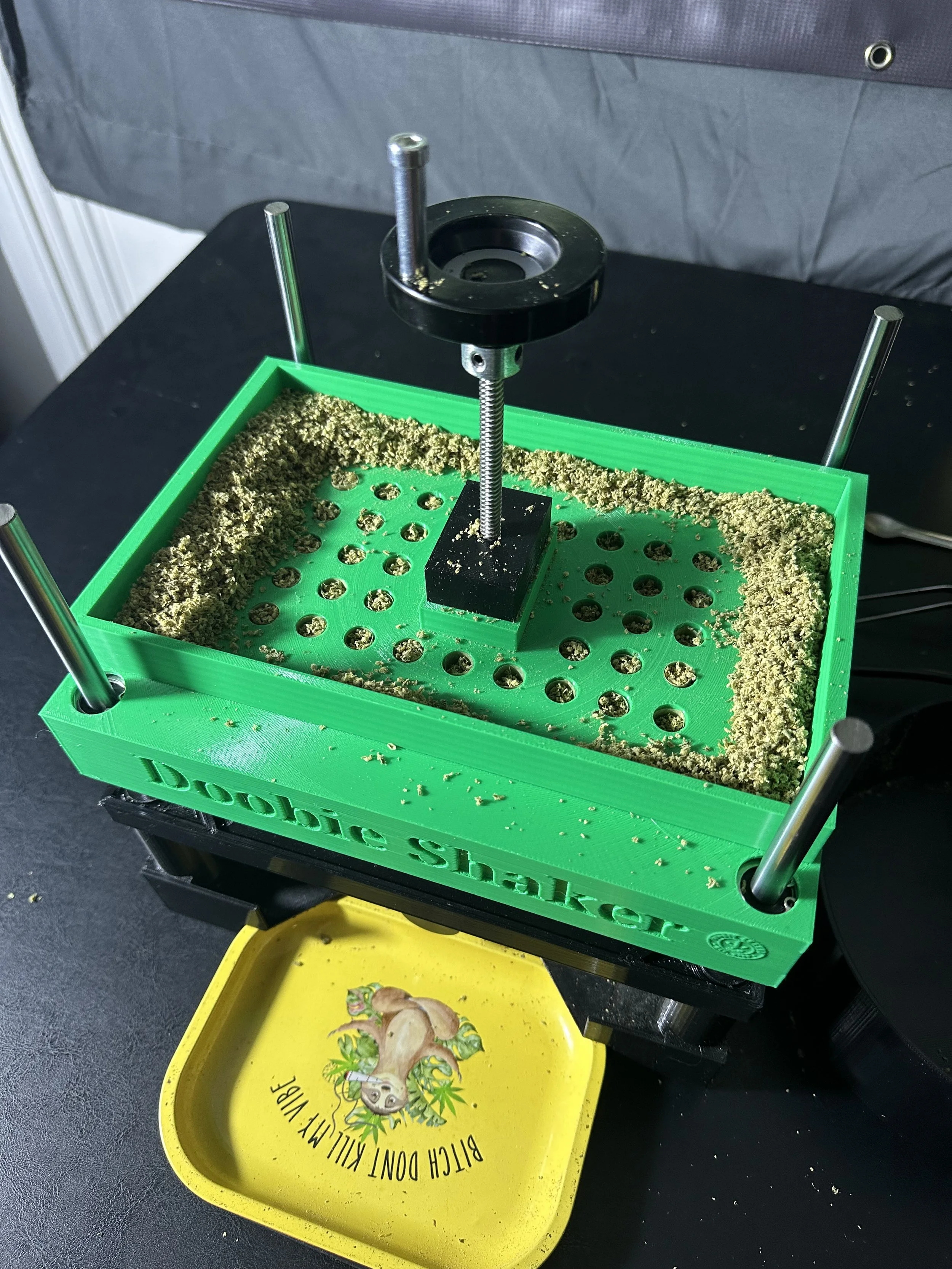 A green plastic device labeled 'Dubble Shucker' with metal posts, round holes, and a central screw mechanism, used to open mollusks, placed on a black surface. A yellow tray with a sloth cartoon and the text 'Bitch dont kill my vibe' is underneath.