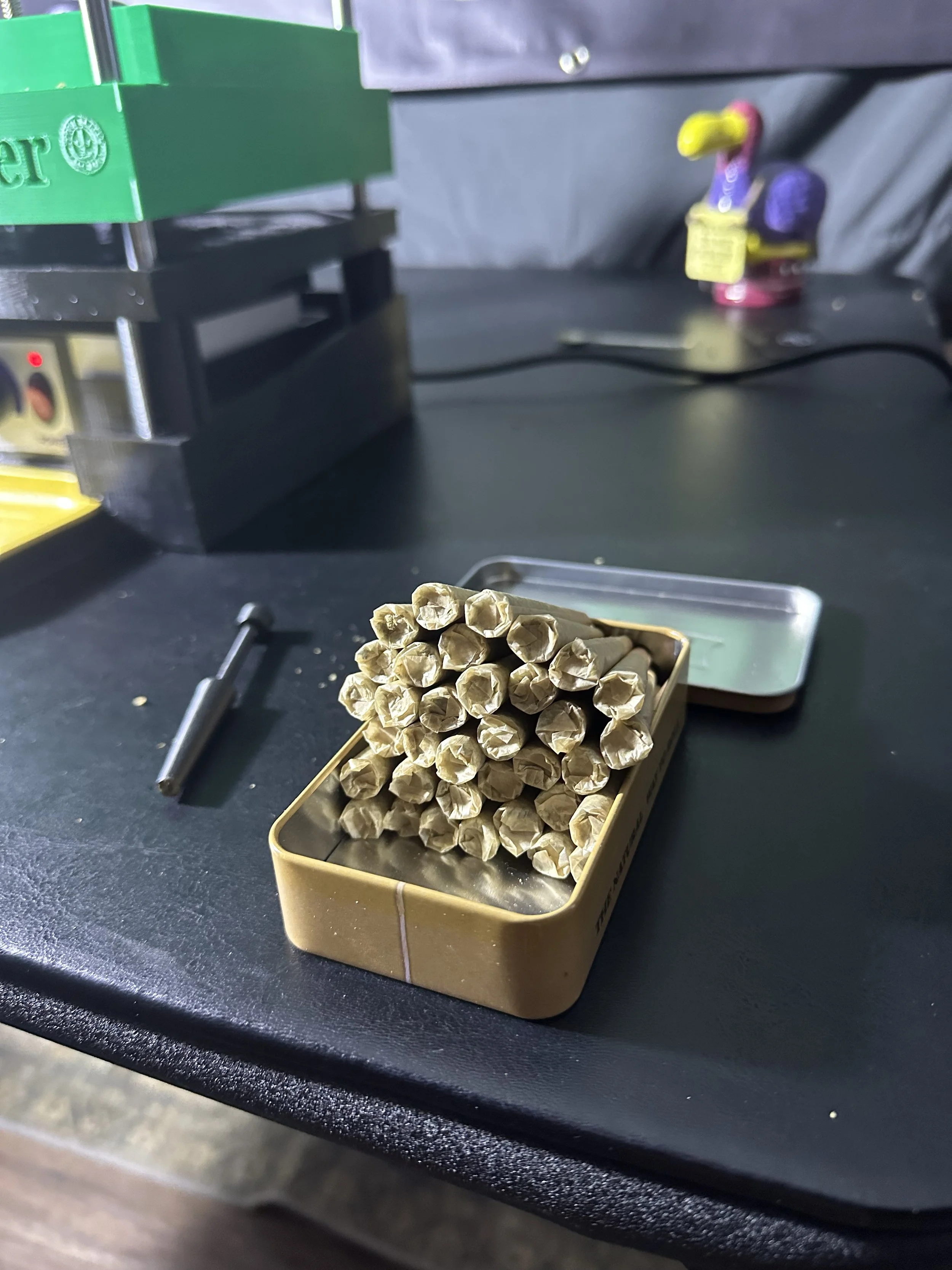 A tin container filled with unlit matches on a dark work surface with a pen and an electronic device in the background.
