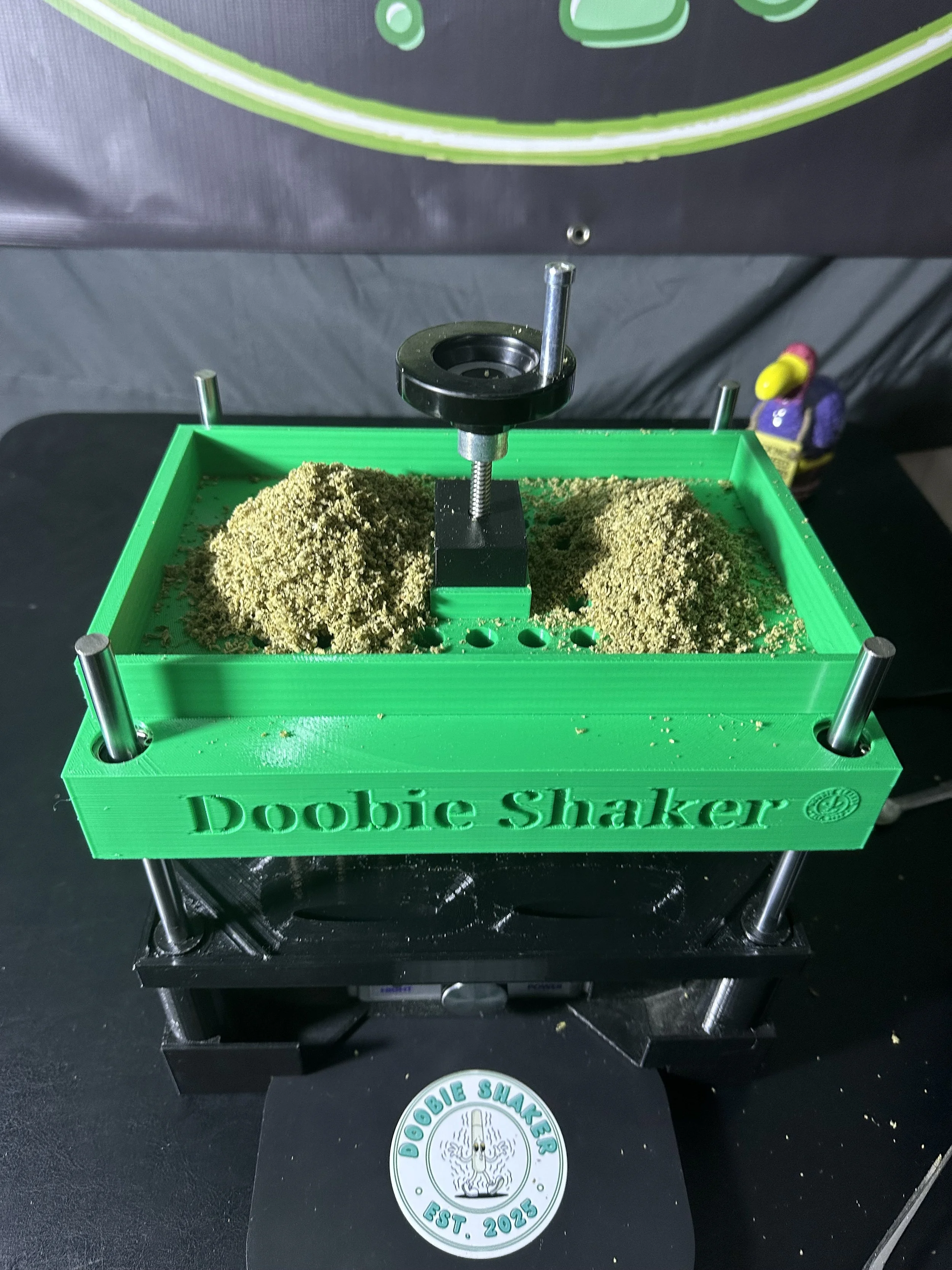 A green Doobie Shaker device filled with ground cannabis, with a black base and metal rods, sitting on a black surface.