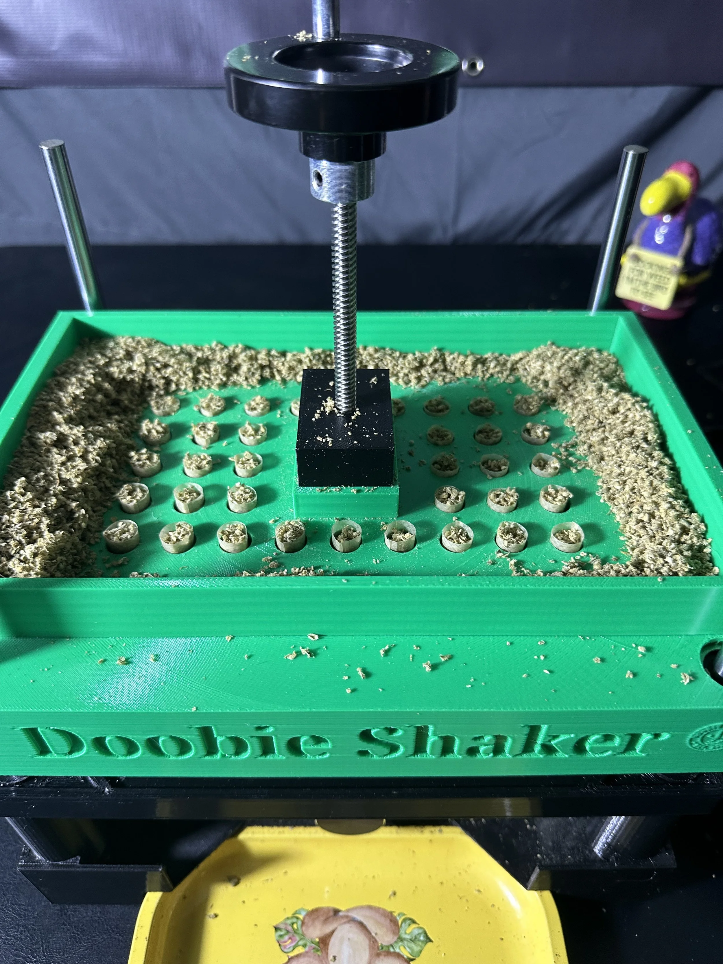 A green plastic tray labeled 'Doobie Shaker' filled with small, round, pre-rolled marijuana cones and covered with ground cannabis. There is a metal shaking mechanism on top, with a black circular handle, used to shake the tray and fill the cones.
