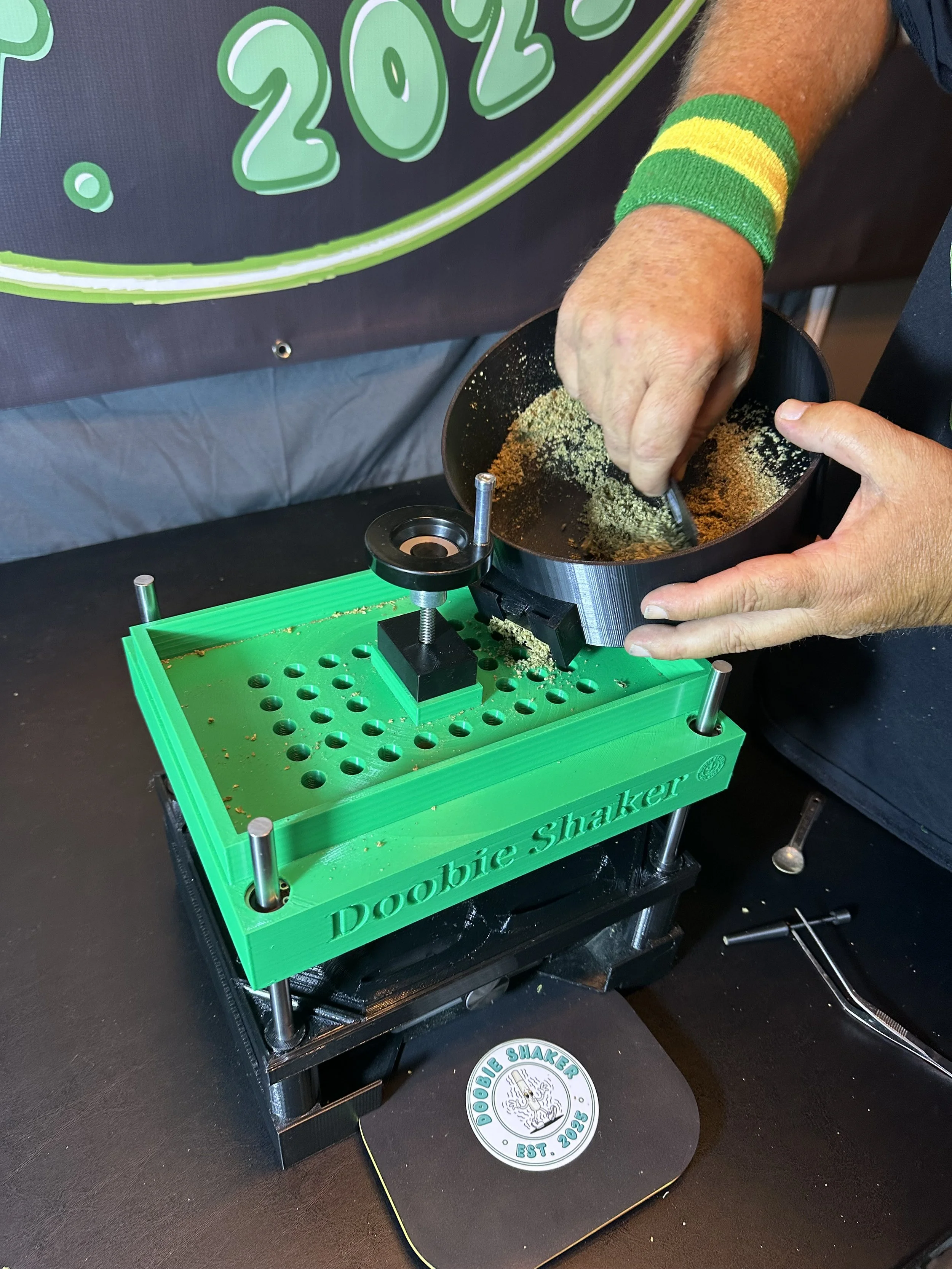 Person using a green 'Dooobie Shaker' machine to mix or grind cannabis or herbs in a black bowl, with tools and a sticker on the table nearby.