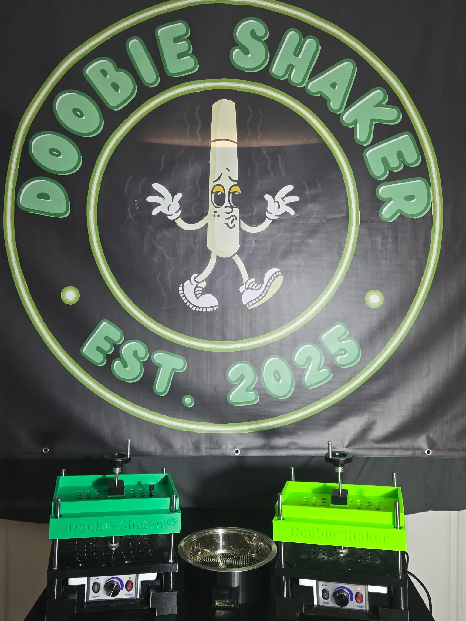A banner with a cartoon cigarette character wearing sneakers and making peace signs, labeled 'Doobie Shaker,' with the text 'EST. 2025' and the name 'Doobie Shaker.' Below are two green shaker devices on tables.