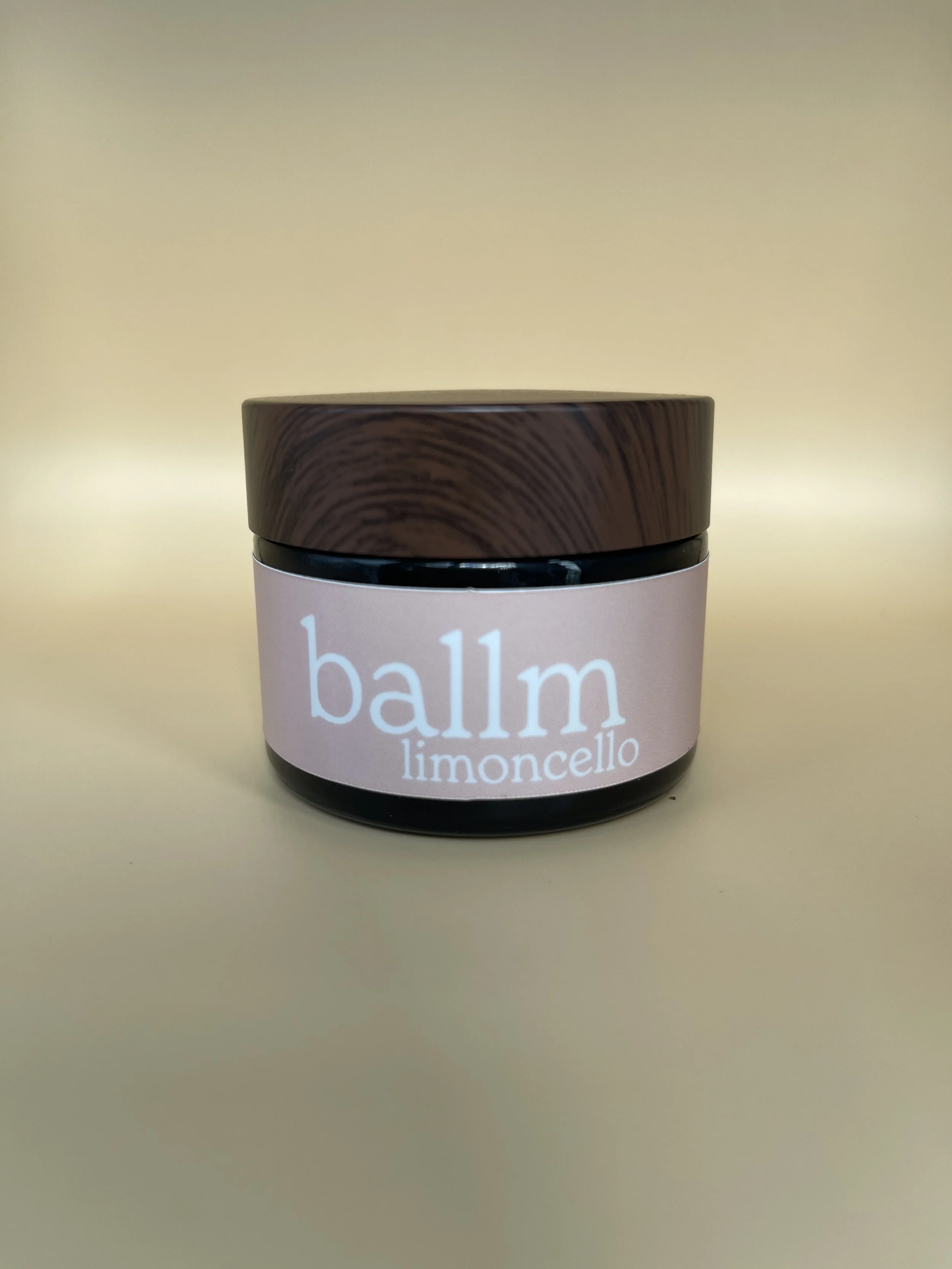 A container of balm labeled 'balm limoncello' with a wooden cap and pink label is placed on a beige surface against a beige background.