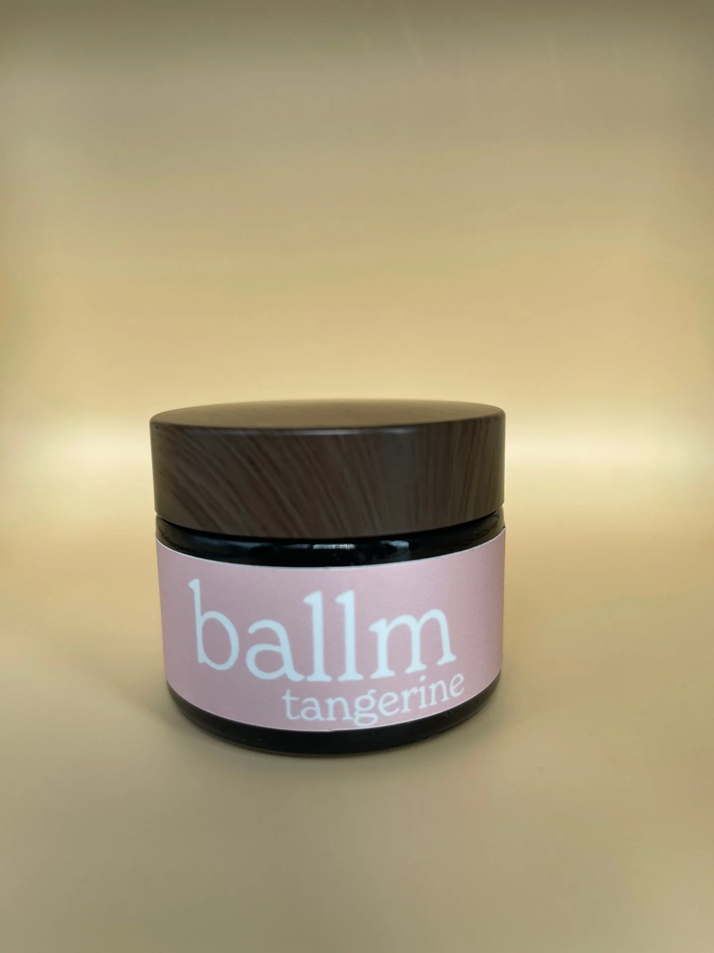 A small jar of lip balm labeled 'balm tangerine' with a dark brown lid, against a plain beige background.