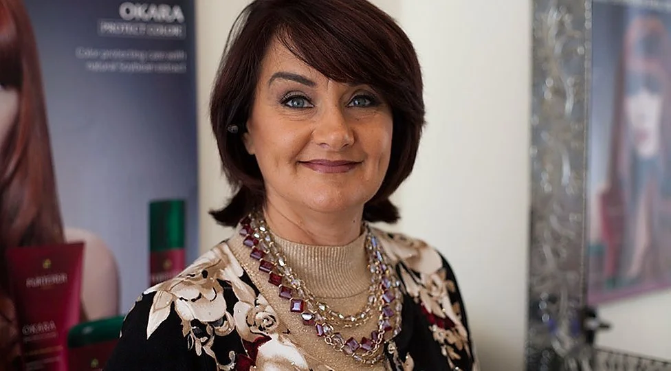 A woman with short brown hair and blue eyes smiling at the camera, wearing a floral top and layered necklaces, standing indoors with promotional posters and decorative mirrors in the background.