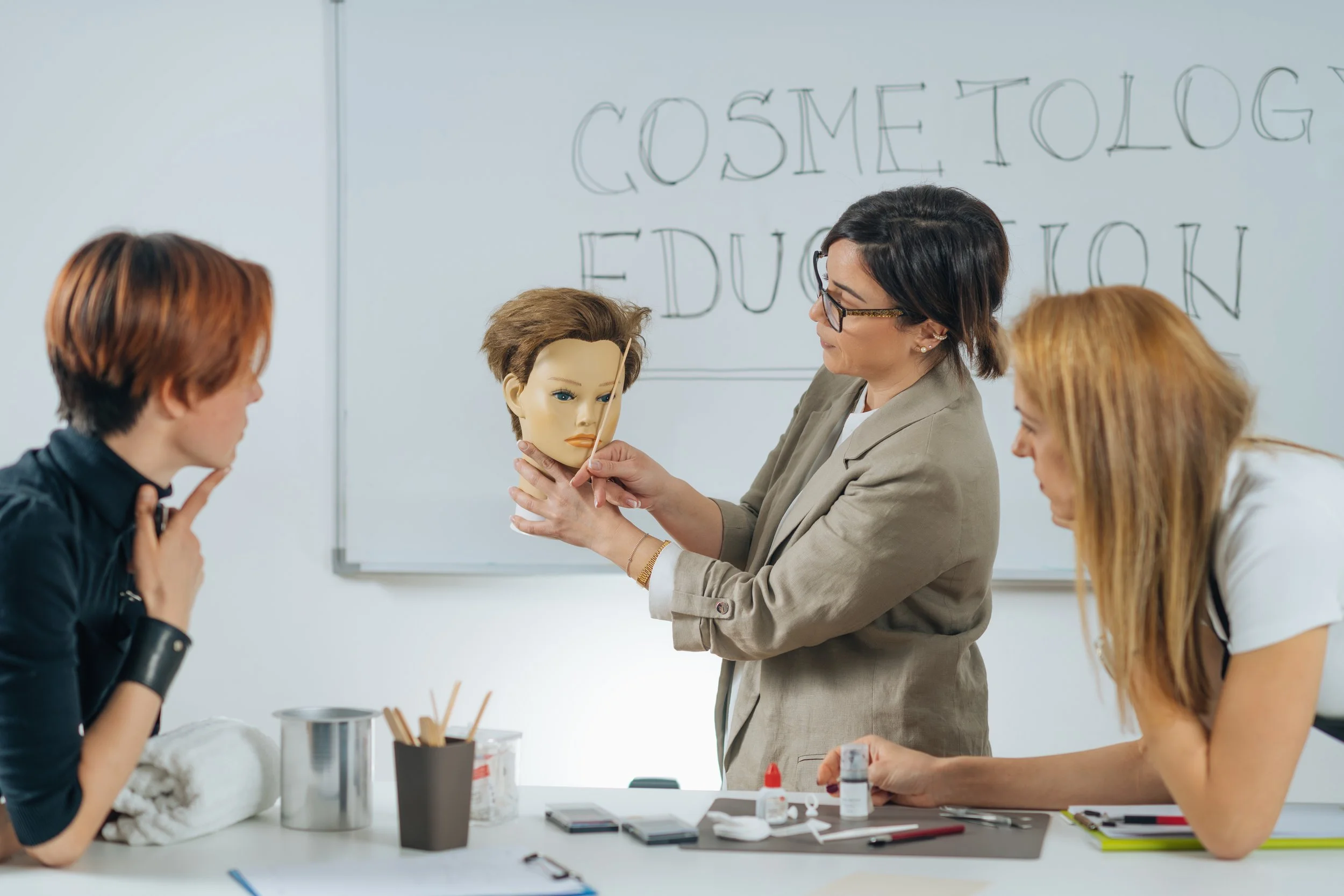 A woman instructor demonstrates how to do a makeup application using a mannequin head in a classroom setting with two students observing.