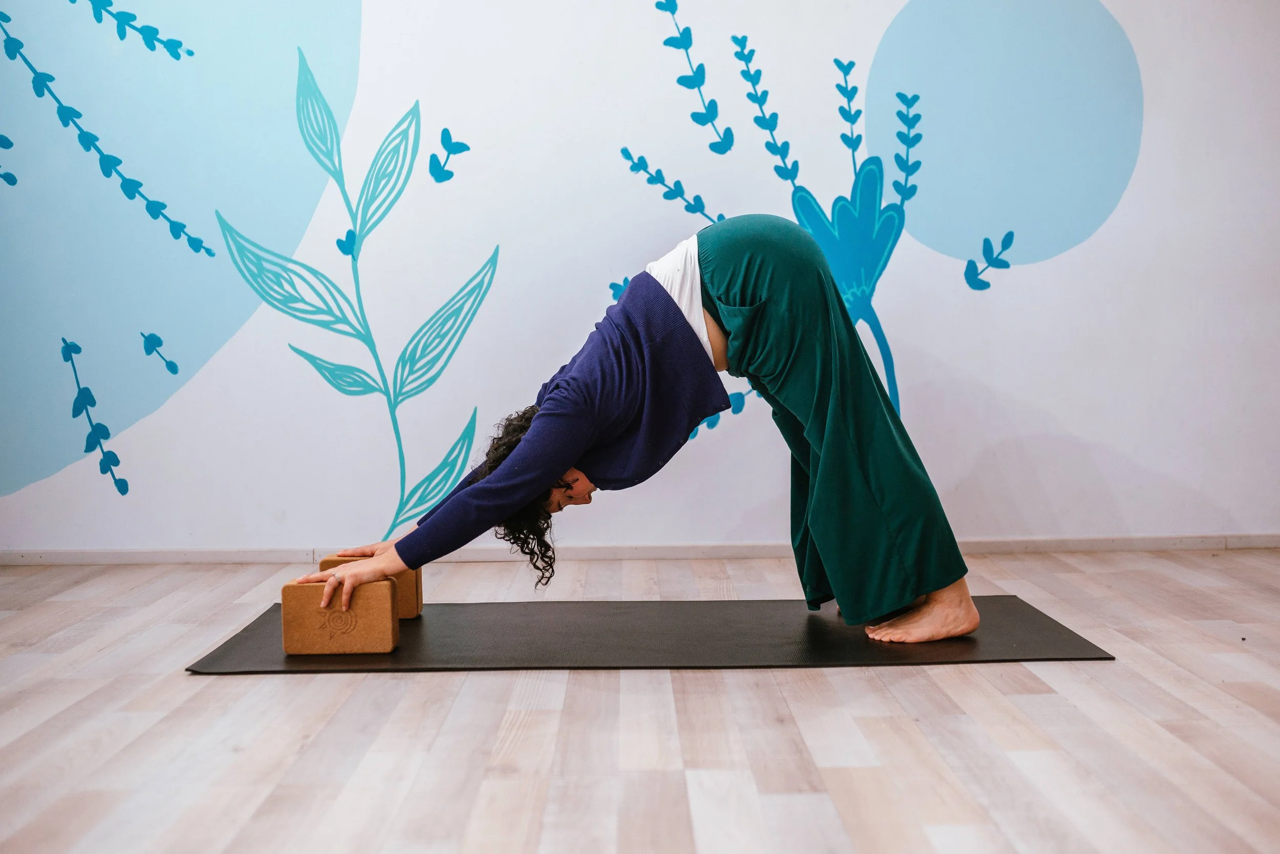 A woman with purple hair and tattoos on her arm practicing aerial yoga using a blue hammock in a studio with blue floral wall decorations.
