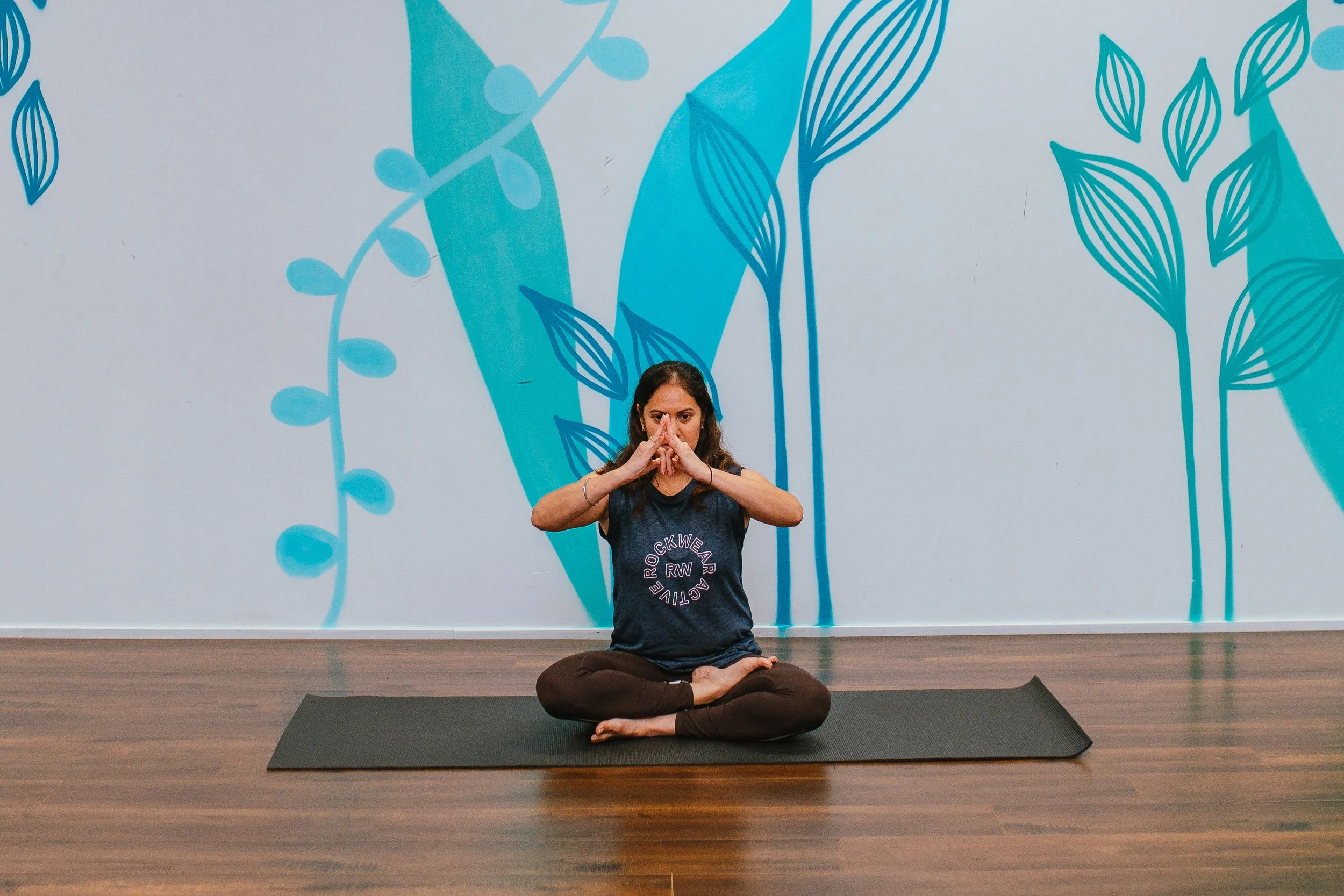 People gathered in a brightly lit room with large windows, sitting on cushions on the wooden floor and talking. In the background, some people are standing near a wall with posters, and a circle of candles and flowers is on the floor.