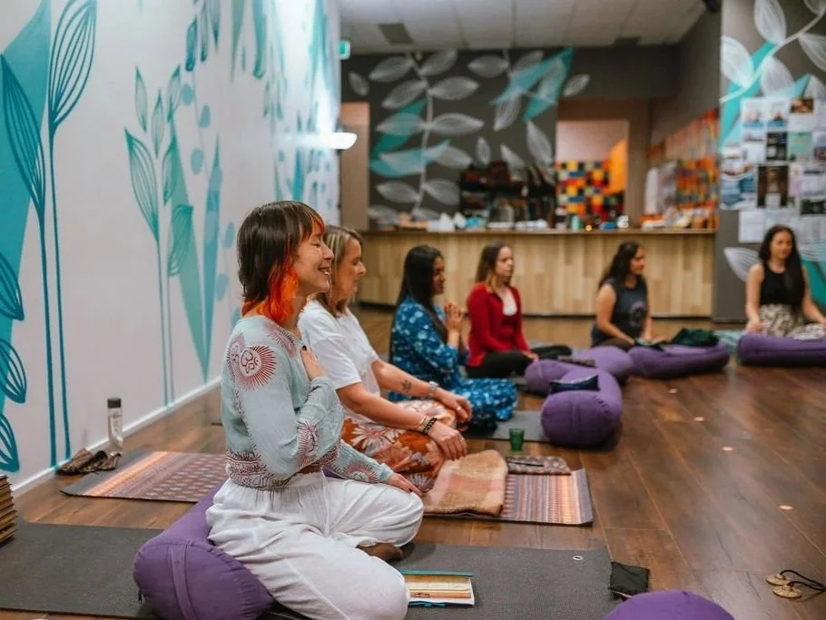 Woman practicing aerial yoga in a studio with white brick walls and wooden beams, hanging from teal fabric hammocks.