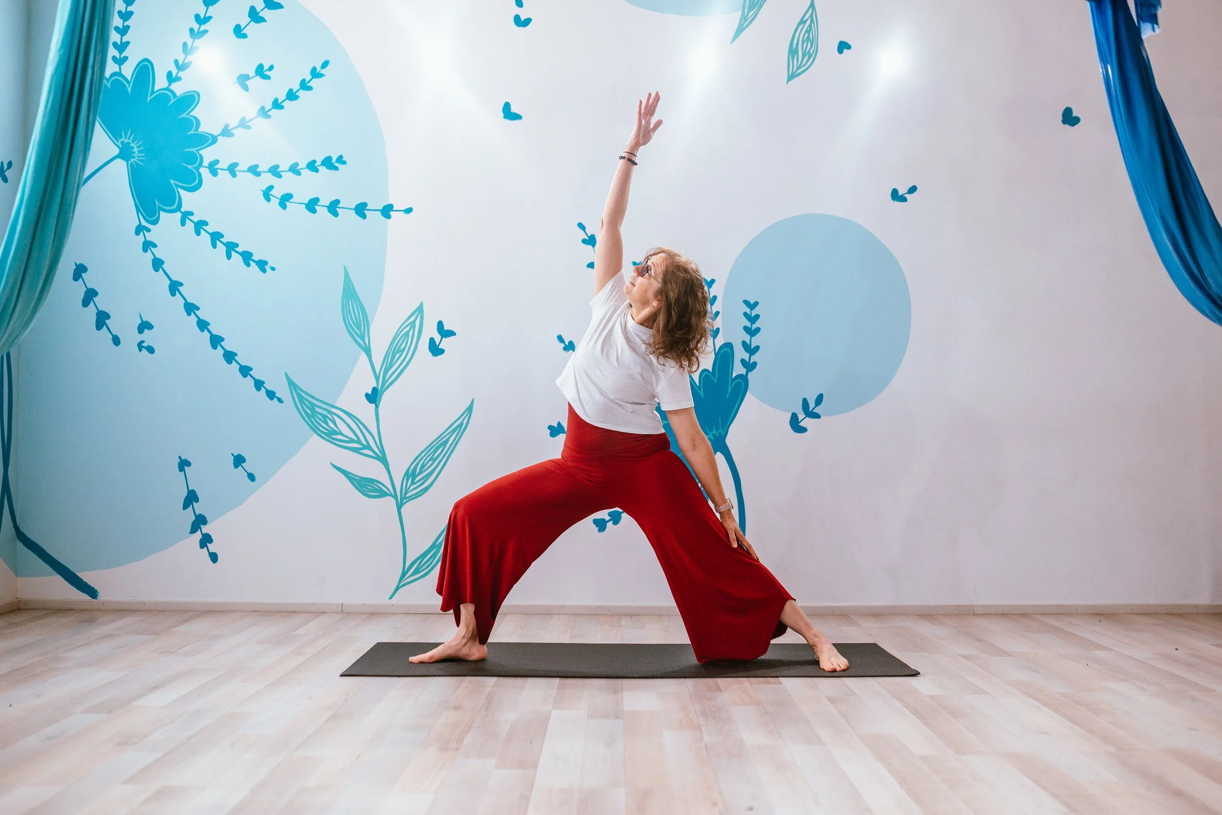 Group of women and girls chatting indoors, with a painting, plants, and shelves in the background.