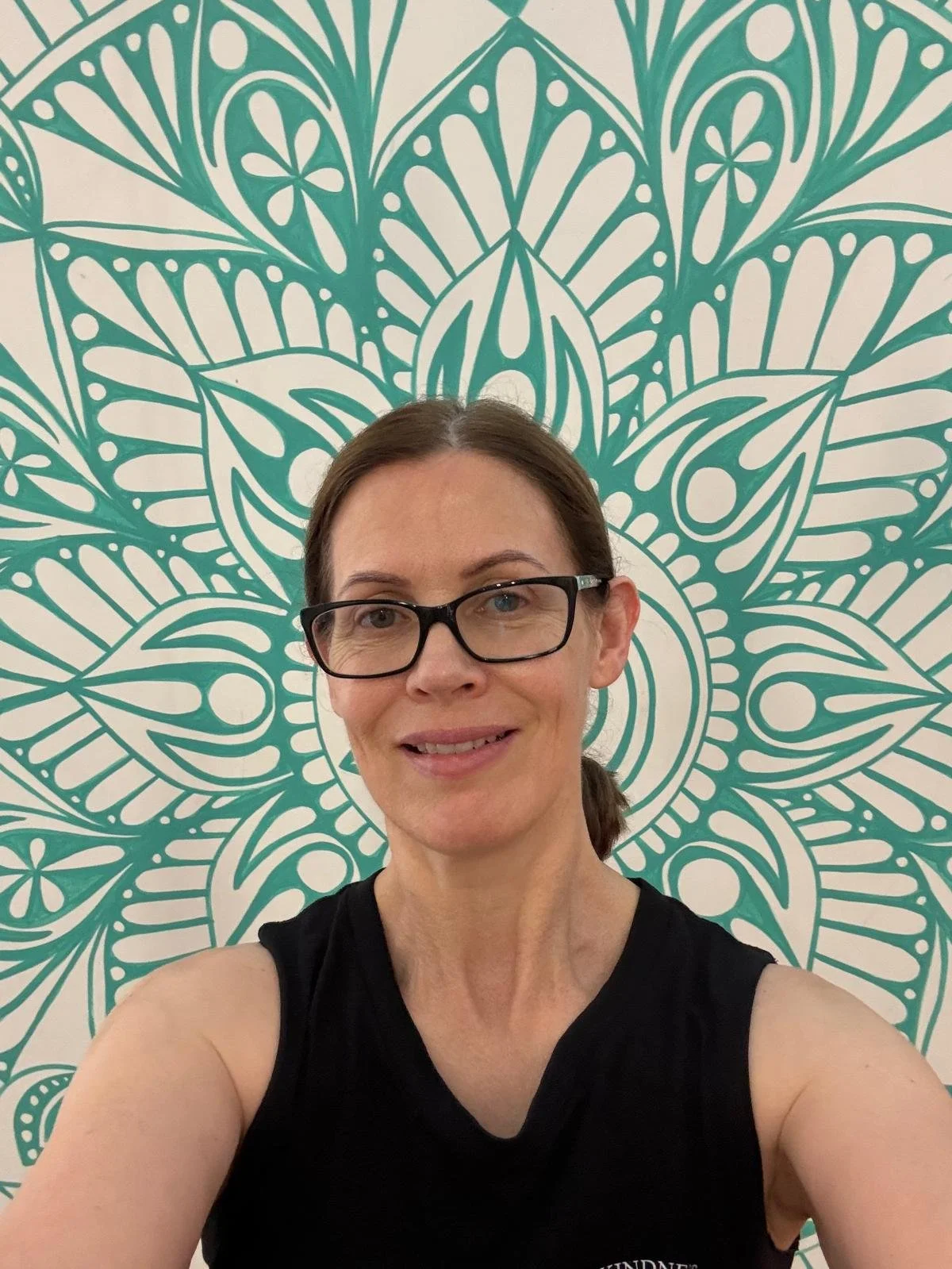 A woman with glasses and a black sleeveless top taking a selfie in front of a decorative teal and white patterned background.