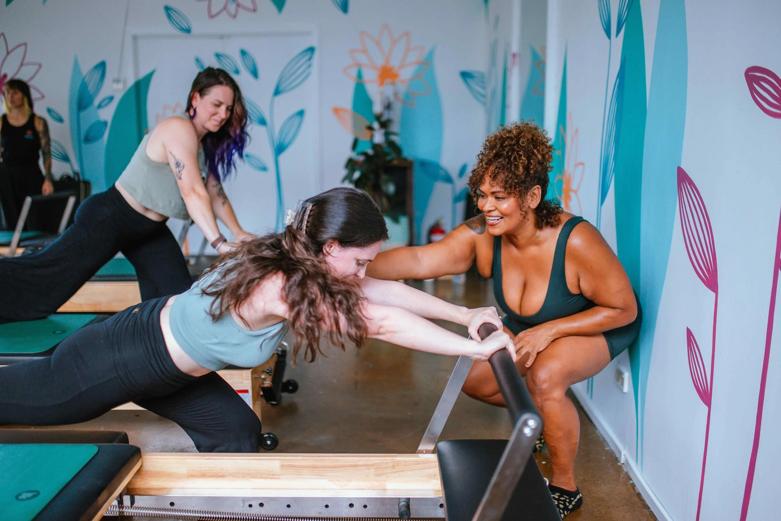 People gathered in a brightly lit room with large windows, sitting on cushions on the wooden floor and talking. In the background, some people are standing near a wall with posters, and a circle of candles and flowers is on the floor.