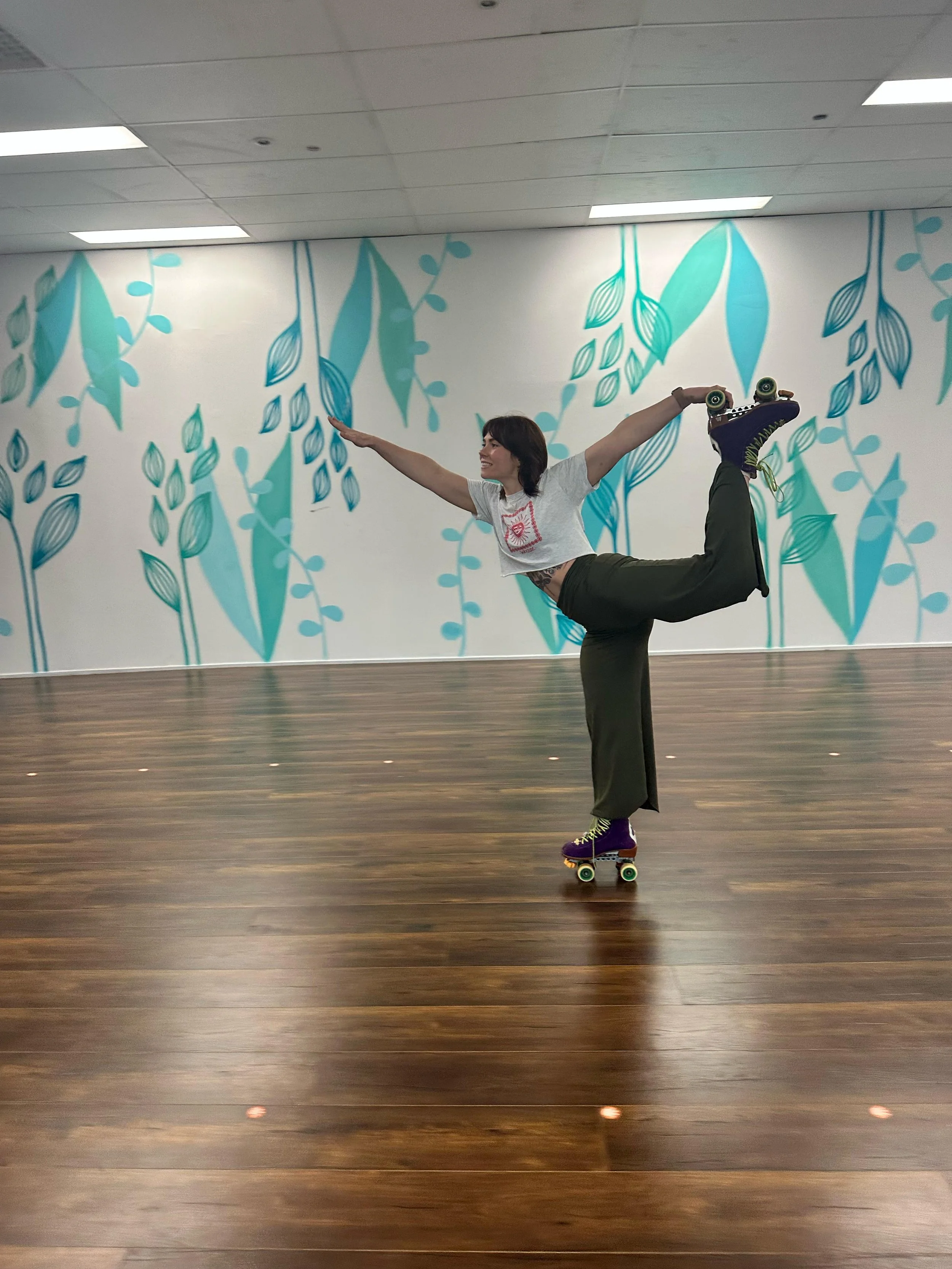A woman smiling while hanging from a green aerial hoop indoors, with colorful mural and geometric wall art in the background.
